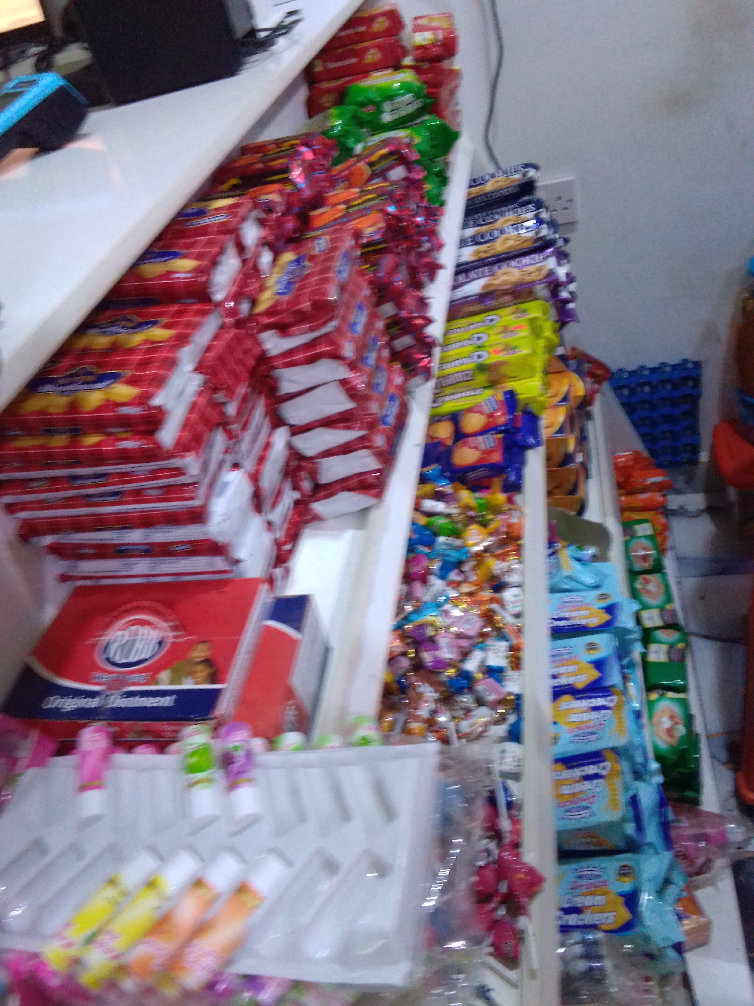 The image depicts the interior of a retail store, likely a small convenience store or a section of a larger market, located in Howayi, Nigeria. The focus is on shelves stocked with various packaged goods, primarily snacks and confectionery. Prominently displayed are stacks of red and yellow packaged biscuits or crackers, as well as green and red packages that appear to be biscuits as well. Further down the shelves, there are packages of cookies, with labels that read "Chocolate Cookies." There is also a display of what appear to be small tubes, possibly lip balm or similar personal care items, in various colors including pink, green, and purple. Beneath these, further rows of colorful candies and wrapped snacks are visible. On a lower shelf, a red box with blue and white lettering, possibly medicine or ointment, is present. In the background, blue plastic crates are stacked, and some loose bags of snacks are also visible. The lighting suggests an indoor environment, possibly with artificial lighting. No people are clearly visible in the frame. The overall impression is one of a well-stocked retail display within a commercial setting in Nigeria.