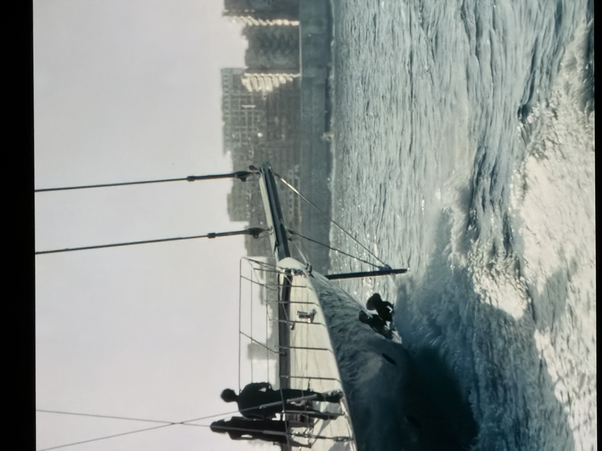 The image shows a submarine moving on the water near the city of London, United Kingdom. Two people are on deck. The buildings of the city are visible in the background. The water is choppy.