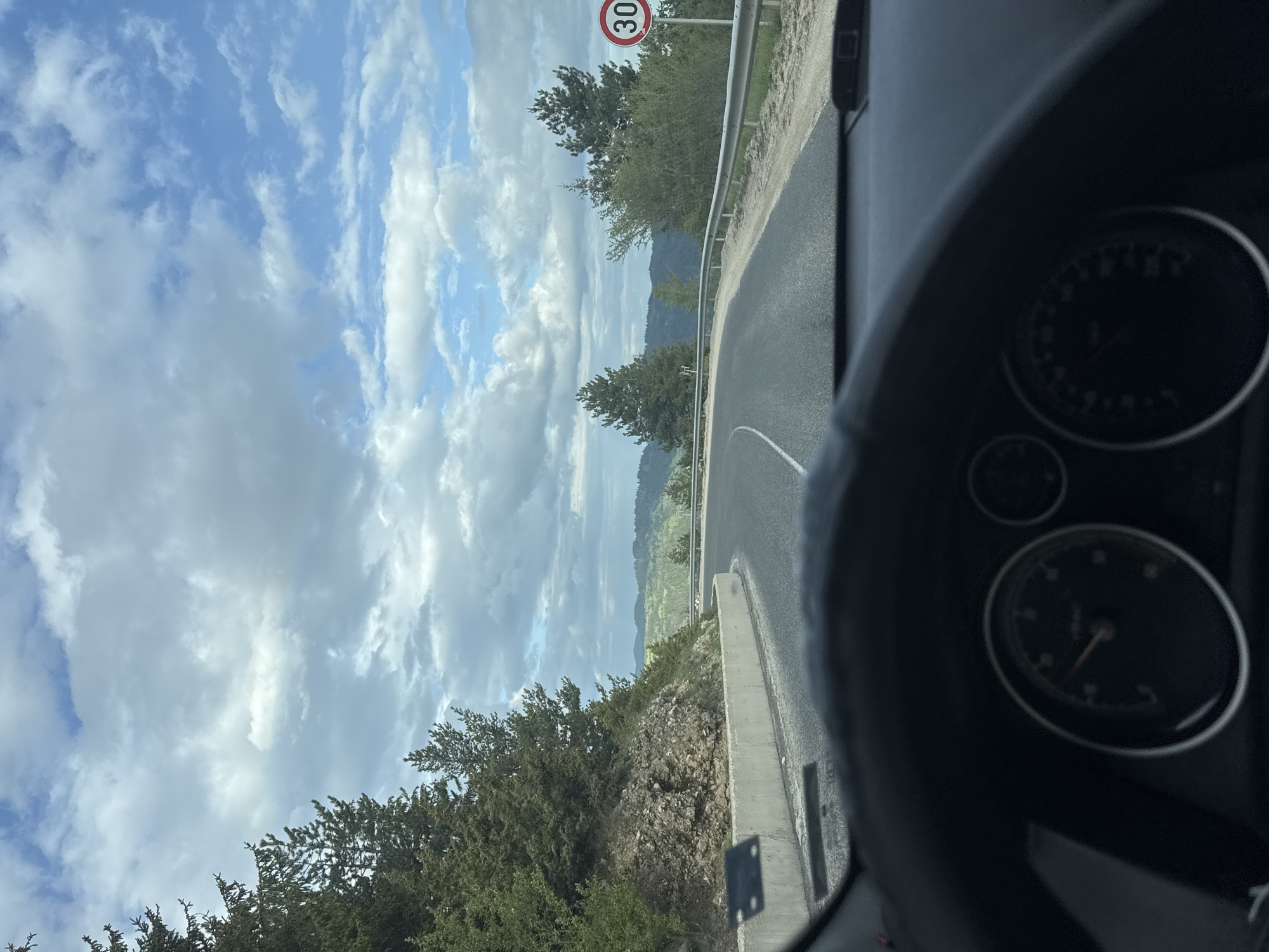 The image depicts a view from inside a vehicle, likely a car, driving on a winding road. The dashboard is partially visible in the foreground, showing gauges. Through the windshield, the road curves to the right, flanked by a guardrail and vegetation. A speed limit sign indicating "30" is visible on the left side of the road. The background features a landscape with trees, hills, and a cloudy sky. The location is Sokolovtsi, Bulgaria, based on the presence of the speed limit sign, which is consistent with European road signage. No people are visible. The focus is on the driving experience and the surrounding landscape.
