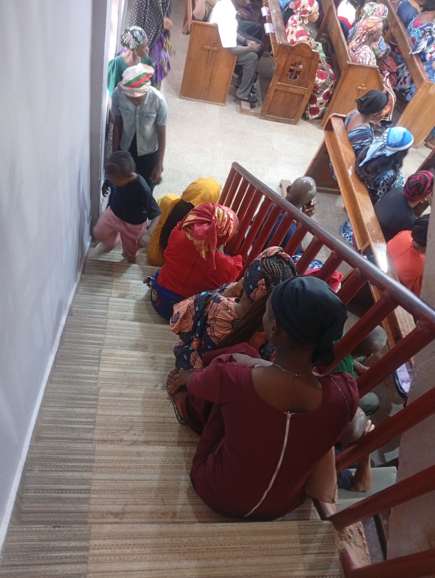 Here is a forensic description of the image:
**Scene:** The image shows an indoor scene, likely a church or similar religious gathering. The location is Unknown, Unknown.
**People:** The image contains multiple people, predominantly women and children. Many are sitting or kneeling on the floor near a staircase. Others are seated in pews in the background. Many of the women are wearing colorful head coverings. One person in the foreground is wearing a burgundy dress and a black headscarf. A child in a black shirt and pink pants is walking towards the left edge of the image.
**Objects:** The dominant objects include pews, a staircase with red-painted railings, and tiled flooring. The pews are made of wood, with cross details on their sides.
**Actions and Interactions:** The people are in various states of prayer or observation. Some are seated, while others are kneeling or standing. Some appear to be entering the space while others are already in the space.
**Environment:** The interior setting appears to be a religious space. The walls are painted a light color, and the flooring is tiled with a patterned design. The lighting is generally dim but appears to be artificial.
