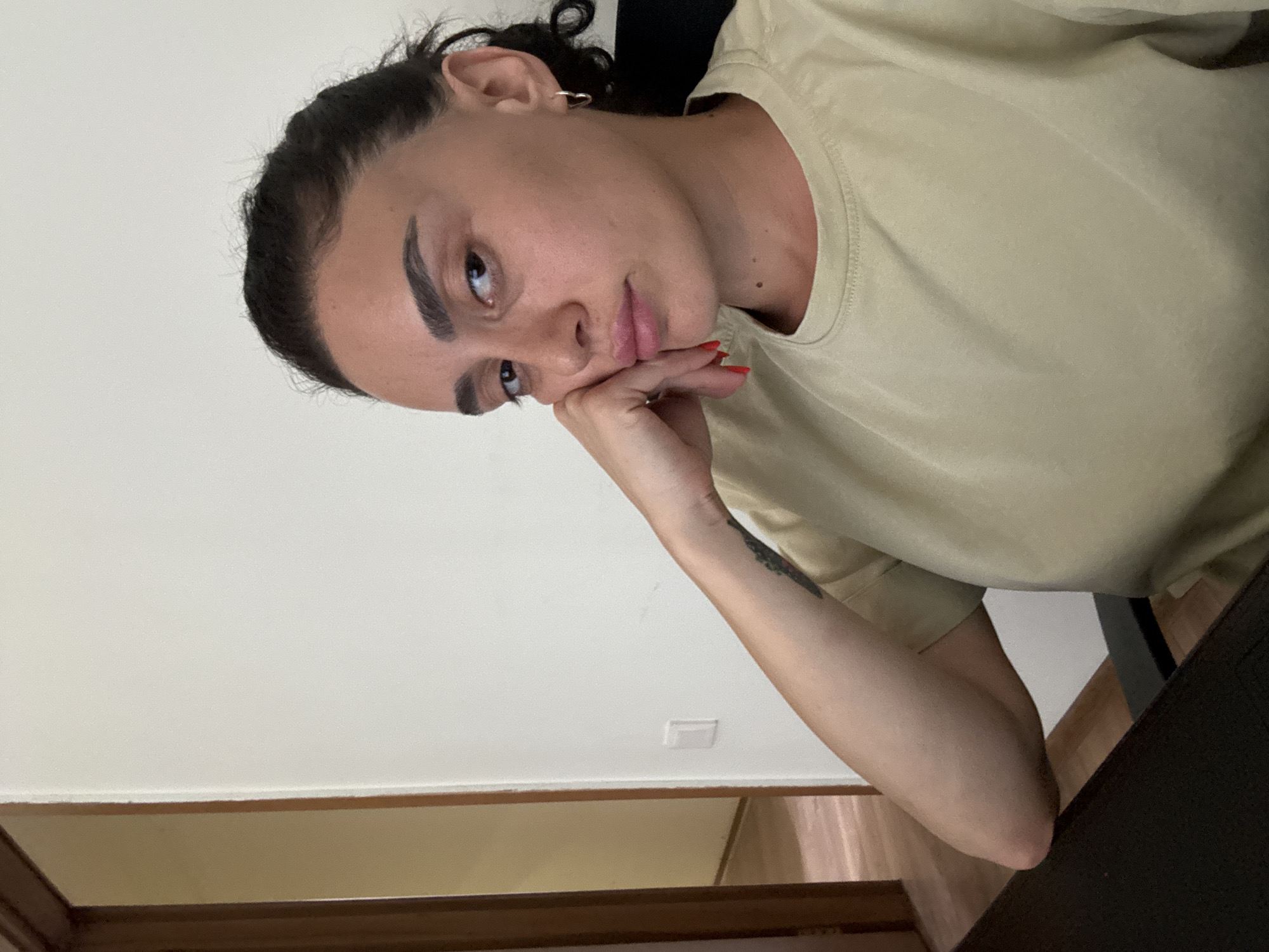 A young woman with dark hair pulled back in a bun is resting her head on her hand, looking at the camera. She is wearing a light yellow t-shirt and has red nail polish. A tattoo is visible on her left arm. She is sitting at a dark desk in front of a white wall. To her left, a doorway leads to a room with light-colored walls and a wooden floor. The scene appears to be indoors, possibly in an apartment or office, located in Tbilisi, Georgia.
