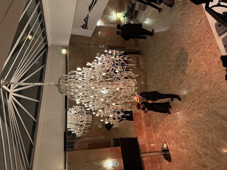 An indoor view from above features a large, ornate chandelier. Below it, a man in a suit and hat stands near a woman in a long dress. The patterned floor reflects the chandelier's light. In the background, other people are visible. Large windows and artwork are positioned near the top of the frame.