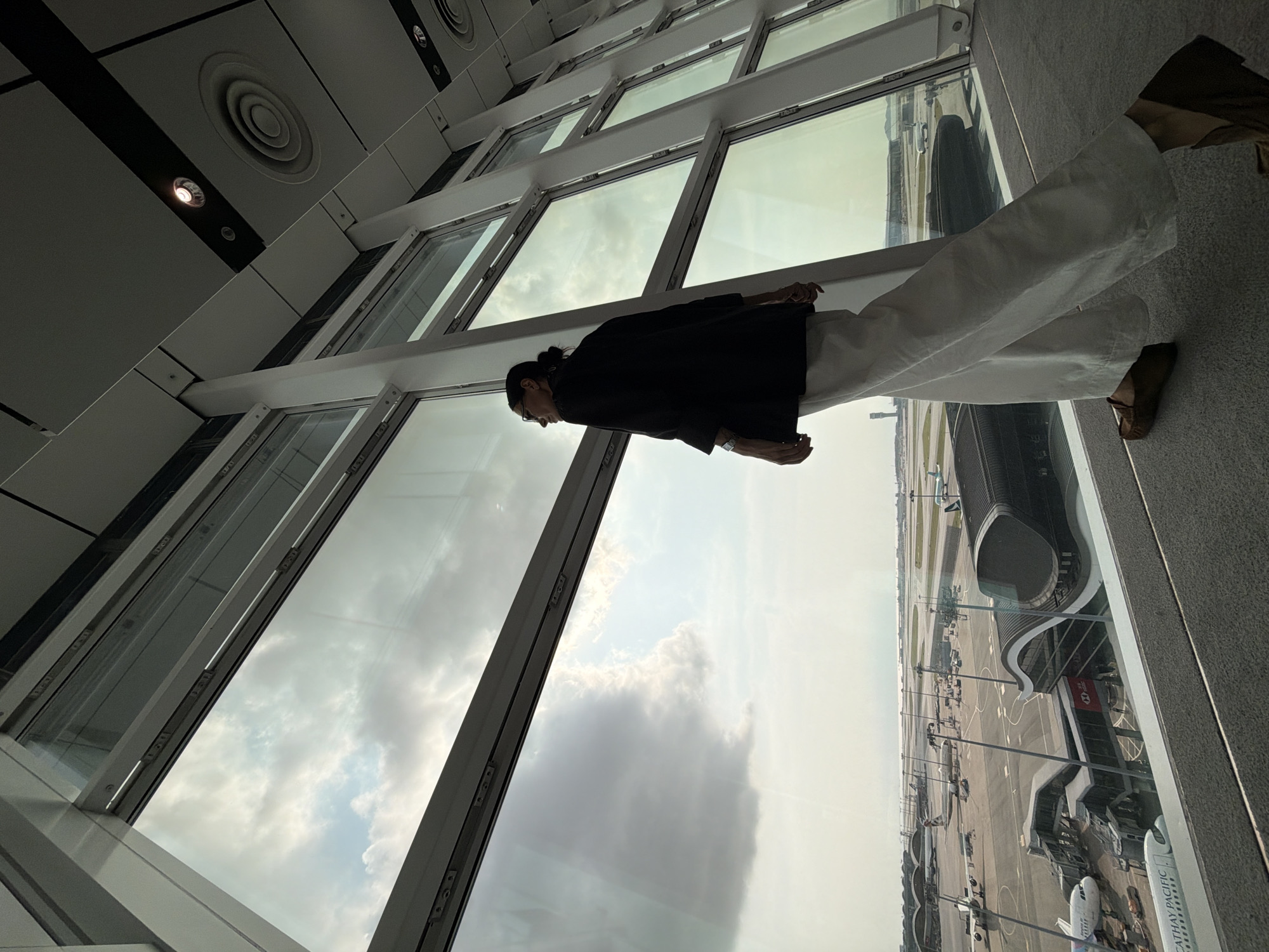 A person stands looking out of a large window at an airport in Hong Kong, China. The person is wearing a black blazer and white pants. The window overlooks an airfield with several airplanes and airport buildings. The sky is cloudy.