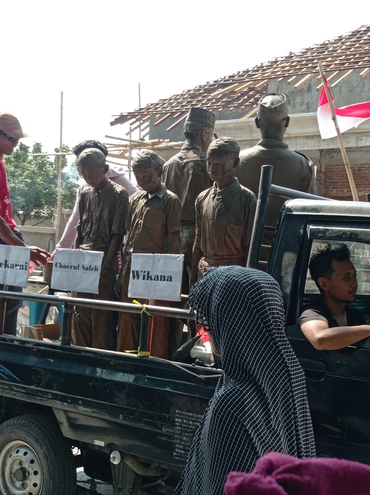 The image depicts an outdoor scene in Maribaya, Indonesia, with a focus on a black pickup truck carrying several mud-covered statues that appear to be of historical figures. The statues are arranged in the truck bed and some have white signs with names written on them. The truck is parked on a dirt or gravel surface. In the foreground, a person wearing a black and white checkered head covering is partially visible, and a person in the driver's seat of the truck is looking to the right. Behind the truck, a building with a partially constructed roof is visible, and a red and white Indonesian flag is flying on a pole. The environment appears sunny and the overall impression is that of a cultural or historical event.