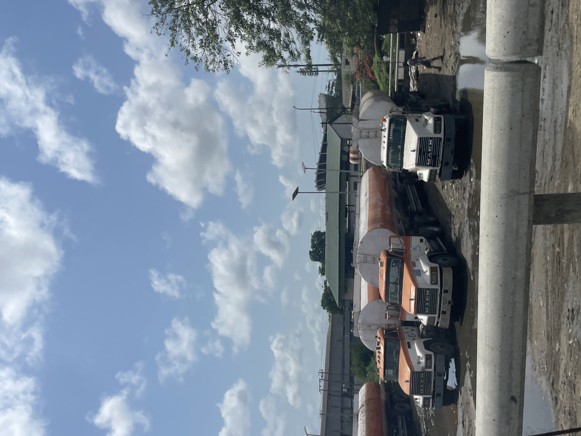 The image depicts several tanker trucks parked in a muddy lot in Alagbede, Nigeria. The trucks are primarily white and orange, with the word "MACK" visible on the front of one. The ground is saturated with water, forming puddles. In the background, there are buildings with green roofs. Overhead power lines are visible, extending into the distance. The sky is blue with scattered white clouds. A person is visible walking in the background. Concrete poles are in the foreground.