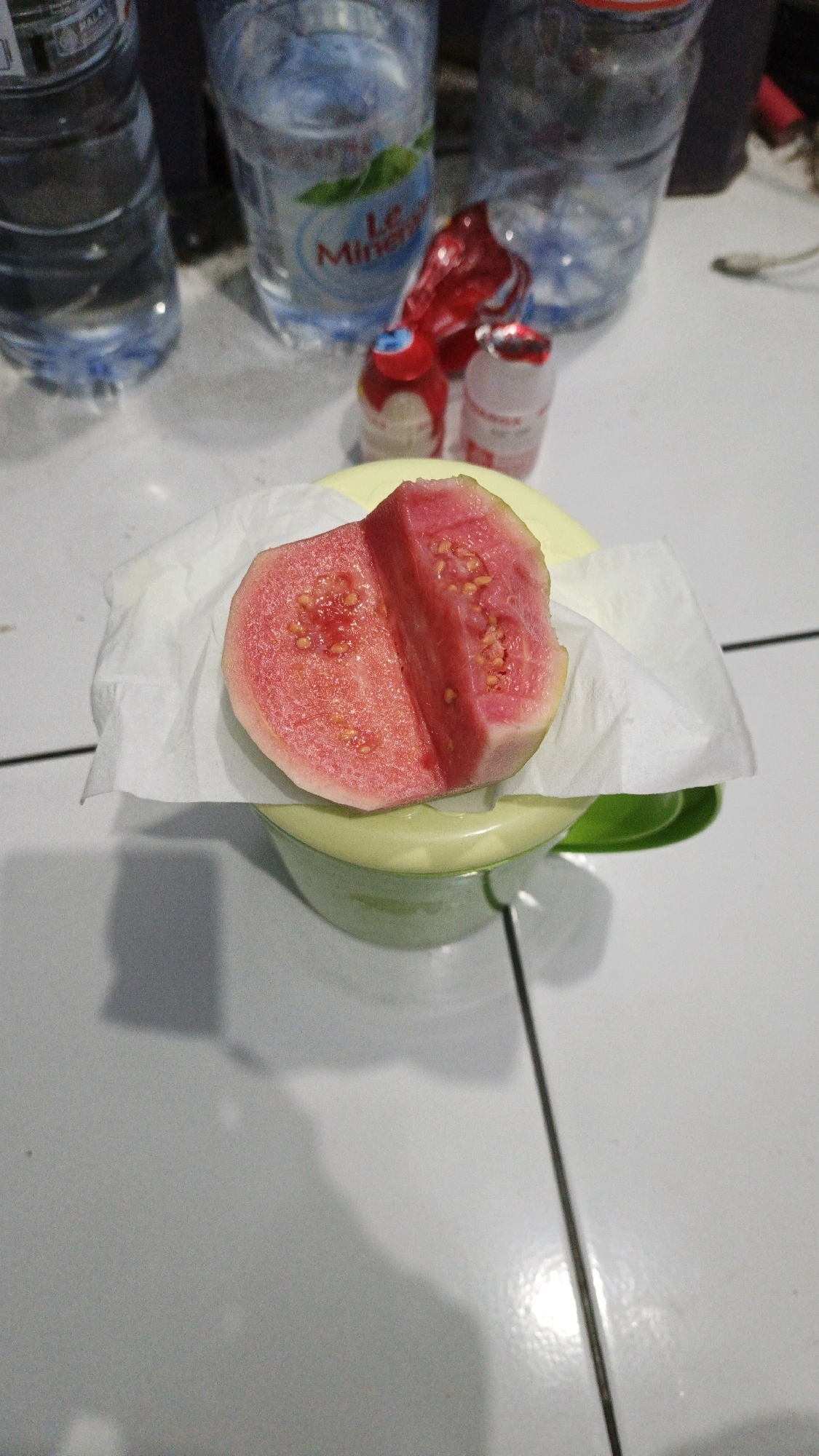 The image displays a close-up, overhead shot of a cut red guava resting on a white paper napkin placed inside a light green bowl. The guava is sliced in half, revealing its pink flesh and numerous small, light-colored seeds. In the background, several plastic water bottles, one with a "Le Minerale" label, and two small red and white plastic containers are visible on a tiled surface. The scene appears to be indoors, possibly in a home or casual dining setting in Campurdarat, Indonesia. The lighting is moderate, casting subtle shadows on the tiled surface.