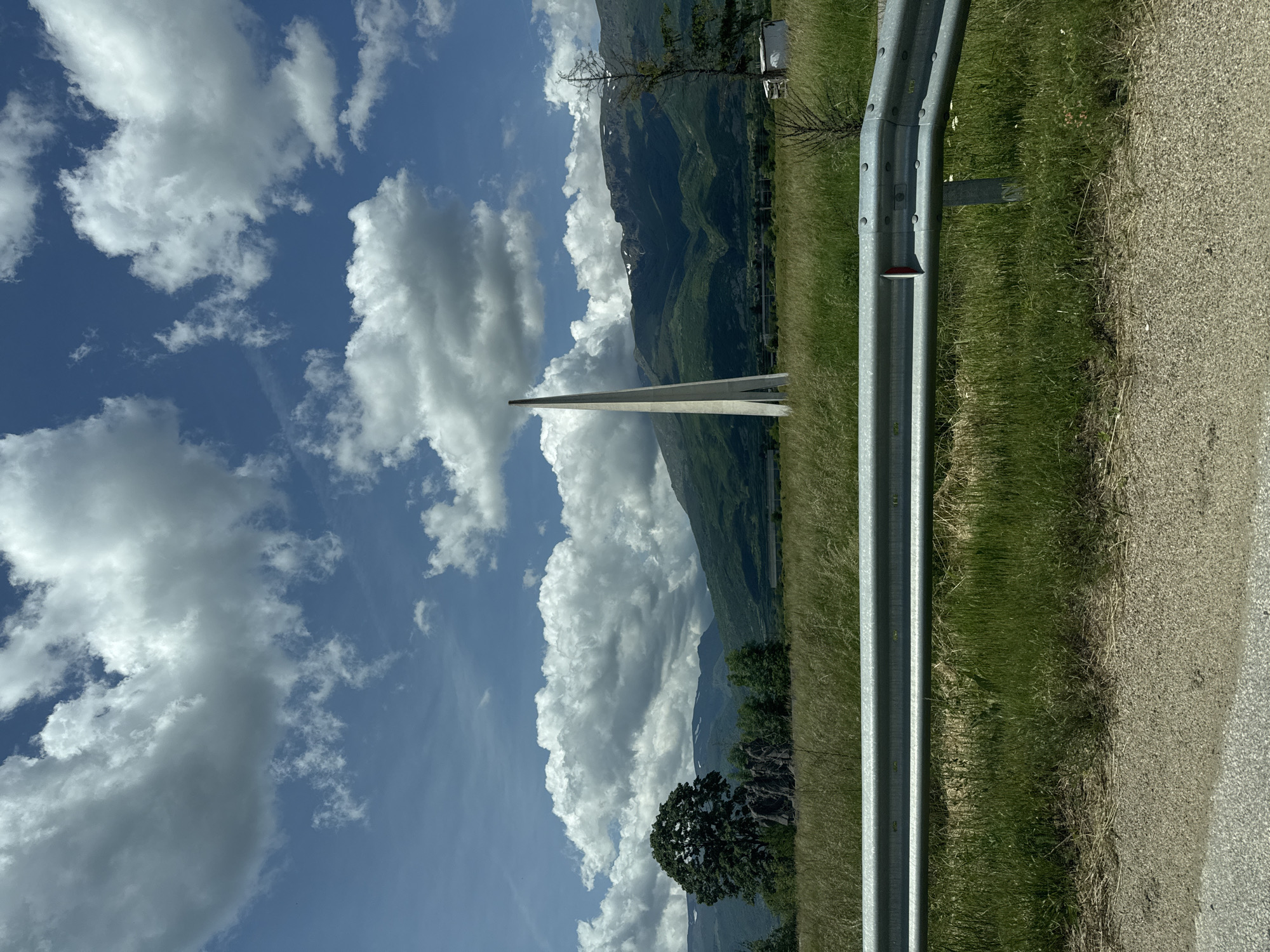 The image shows a landscape featuring a mountain range in the background, partially obscured by large cumulus clouds under a blue sky. A tall, white, modern structure, possibly a bridge support, extends upward in the middle ground. In the foreground, there is a metal guardrail along the edge of a road with green grass and vegetation on the other side of the rail. The location appears to be near a road passing through a mountainous region, but no specific city can be identified.