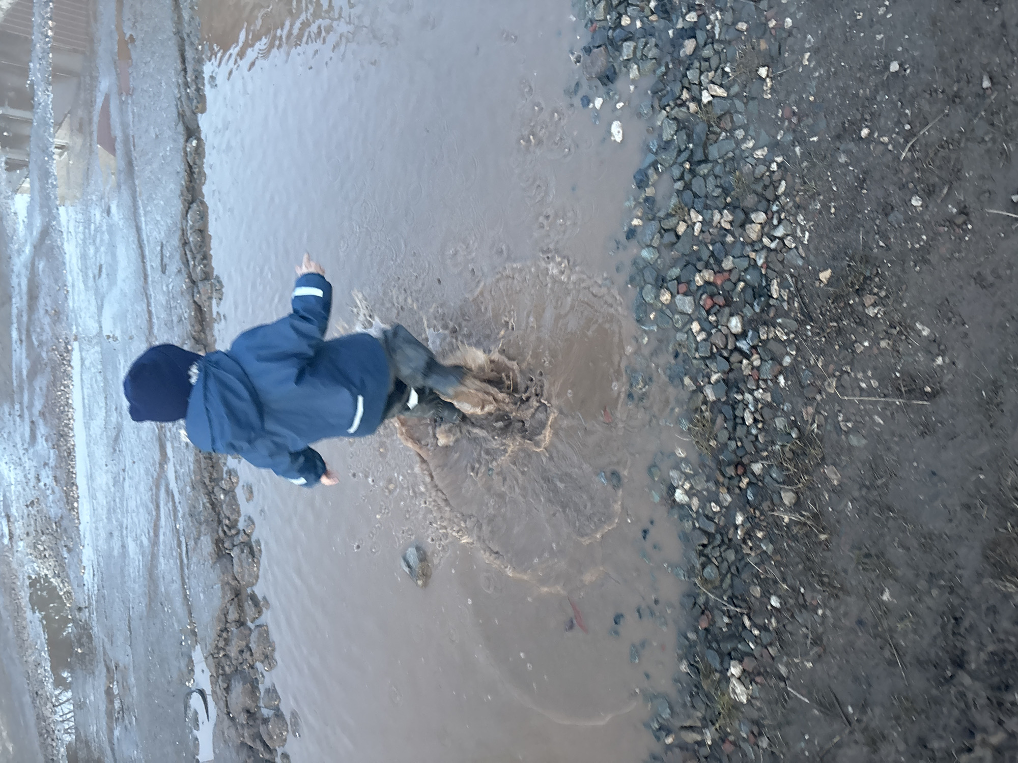A child wearing a dark blue hooded jacket, dark pants, and dark boots is jumping in a large puddle. The puddle is located on a surface that transitions from pavement on the left to gravel on the right. Splashes of water surround the child, indicating movement and impact. The image appears to be taken from above. Ferma, Russia is not discernable from the provided image.