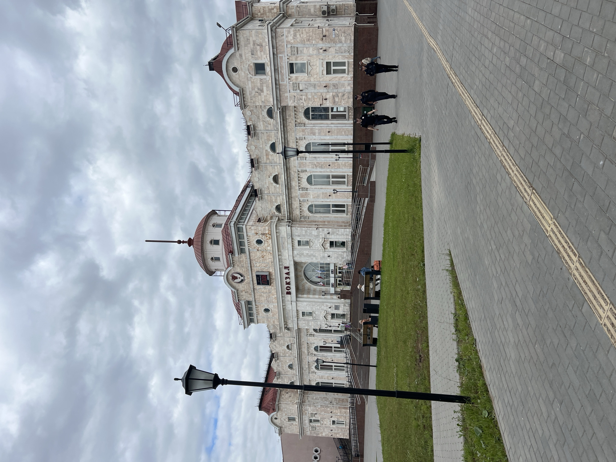 The image shows the Izhevsk railway station in Izhevsk, Russia, on an overcast day. The station is a large, ornate building with a red dome and a clock on its facade. The word "ВОКЗАЛ" (VOKZAL, meaning "station" in Russian) is prominently displayed above the main entrance. In the foreground, a black lamppost stands on the left side of the image. A paved walkway with a tactile paving strip runs diagonally across the lower part of the frame. To the left of the walkway is a patch of green grass.
Several people are visible in the image. Three individuals in dark uniforms, likely police officers, are walking on the paved walkway from right to left. Two individuals are seated on a bench on the grass in front of the station, and a few other people can be seen walking up the stairs towards the station entrance. There are also some red patterned bags placed on the ground near the bench. The sky is heavily clouded, suggesting a temperate or cool climate. The architecture of the station is characterized by classical elements, including columns and arched windows. The building appears to be made of light-colored stone or marble. An air conditioning unit is visible on the right side of the station building.