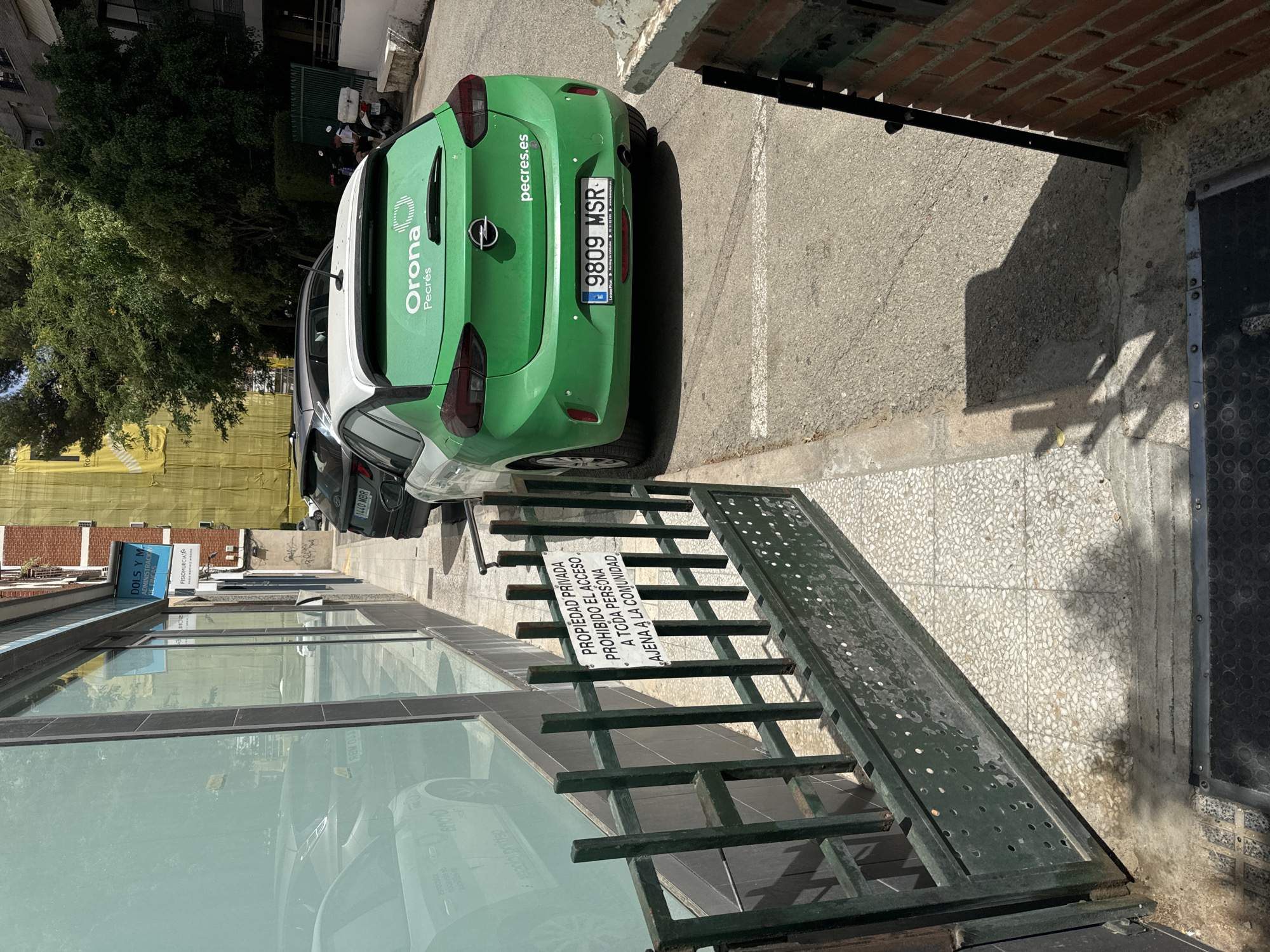 This image shows a street scene in Murcia, Spain. In the foreground, a green Opel car with the branding "Orona Pecrés" and the website "pecres.es" is parked. The license plate reads "9809 MSR". Next to the car, there is a sign that says "PROPIEDAD PRIVADA PROHIBIDO EL ACCESO A TODA PERSONA AJENA A LA COMUNIDAD" which translates to "PRIVATE PROPERTY ACCESS PROHIBITED TO ANY PERSON NOT BELONGING TO THE COMMUNITY". Behind the car, a building with brick elements and trees can be seen. A metal fence with a ramp is also visible in the foreground, with the reflection of buildings in the glass windows. In the background, there are signs for "DOLS Y M ADMINISTRACIO DE FINCAS" and "FISIOMURCIA". The lighting suggests it is daytime, likely sunny. No people are clearly visible in the image.
---
This description is factually accurate and concise, adhering to the requested word count and content guidelines.
