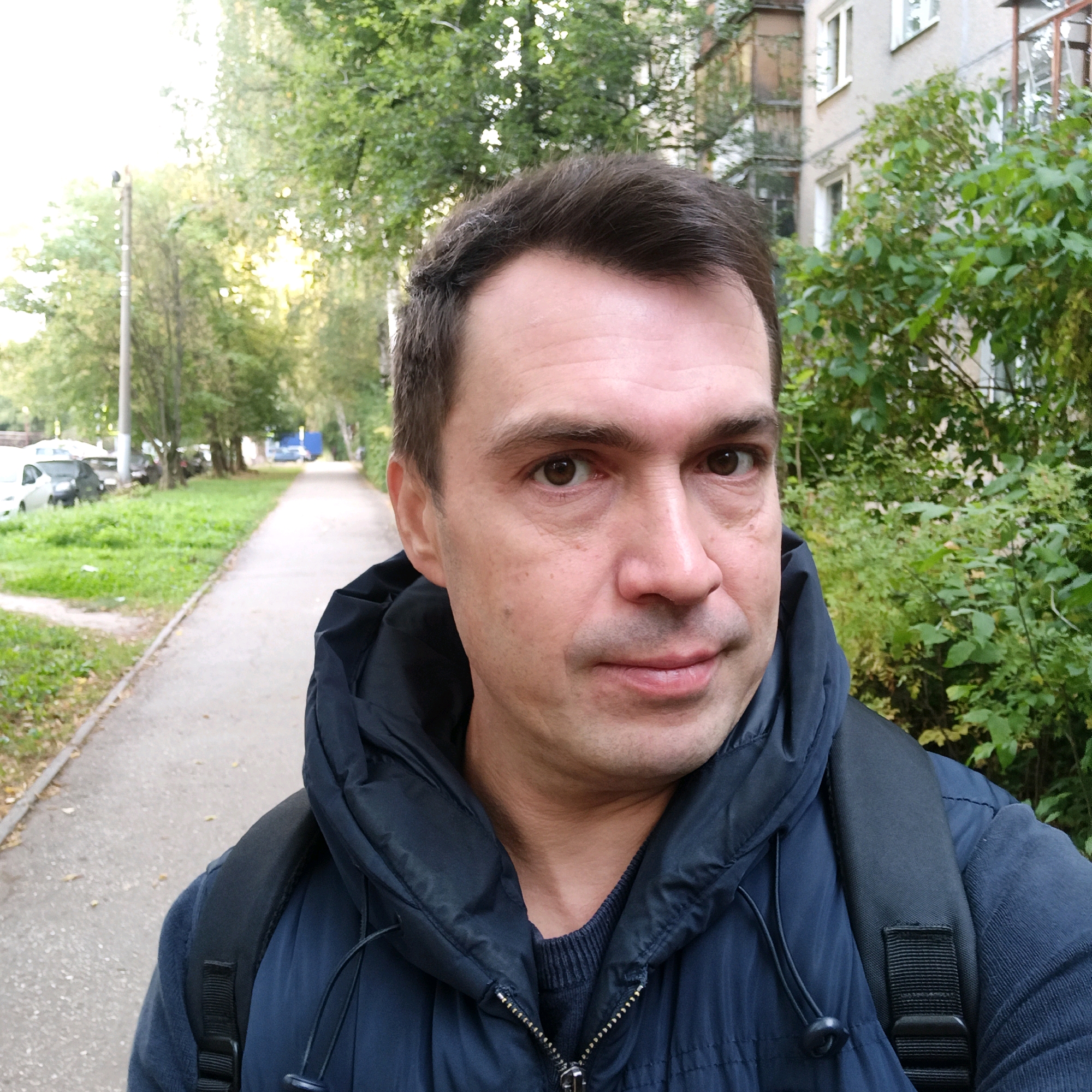 An outdoor, eye-level, selfie-style photograph shows a man in his late 30s to early 40s standing on a sidewalk in what appears to be a residential area of Nizhny Novgorod, Russia. The man has short, dark brown hair, brown eyes, and a fair complexion. He is wearing a dark blue, hooded puffer jacket over a dark sweater. A black backpack is visible on his shoulders. The background consists of a sidewalk, green grass, trees with green and yellowing leaves, and a light-colored building with visible windows and balconies. The lighting suggests it is daytime, possibly late afternoon, with sunlight filtering through the trees. Cars are parked along the side of the street in the distance. The image is taken at an angle that shows the man's face in the foreground and the surroundings in the background.