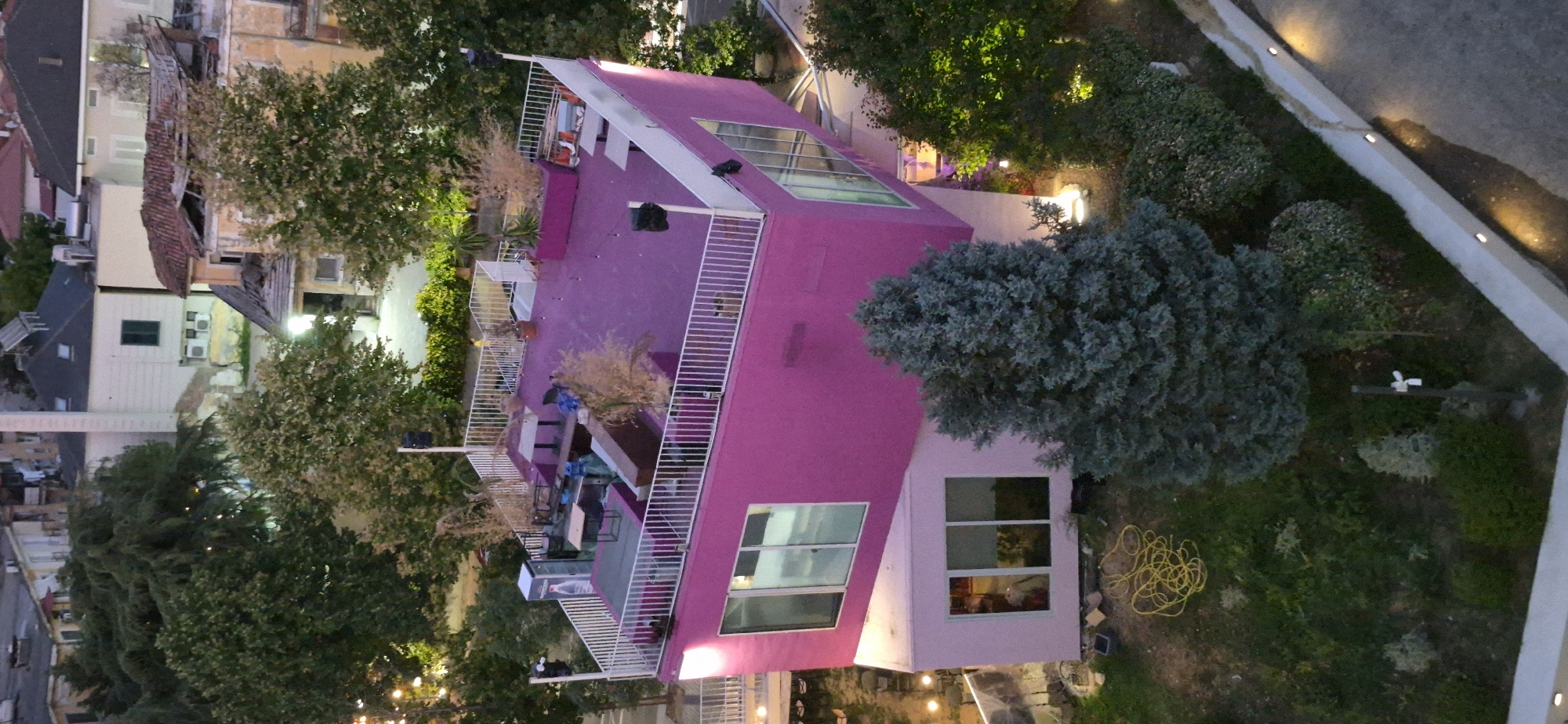 This aerial, high-angle view captures a modern, brightly colored building in Tirana, Albania. The main structure is painted a vibrant magenta, with a contrasting lighter pink lower section. The building features large glass windows. The upper level of the magenta section is topped with a rooftop terrace that has a purple floor. This terrace is furnished with tables, chairs, and planters containing sparse, dry vegetation, suggesting it's set up for outdoor seating or a lounge area. White railings surround the terrace.
Below the magenta structure, a smaller, angled pink section of the building has windows that reflect the interior. In the foreground, a large, dark green coniferous tree stands prominently. To the left of the building, a yellow coiled hose lies on the grassy ground, along with some cardboard boxes, indicating ongoing work or storage. The surrounding environment includes more trees and vegetation, and in the background, other buildings and rooftops typical of an urban setting are visible. The scene appears to be in the evening or dusk, as some artificial lights are illuminated, casting a warm glow on the surroundings. The architectural style suggests a contemporary design that stands out against the more traditional structures in the background.