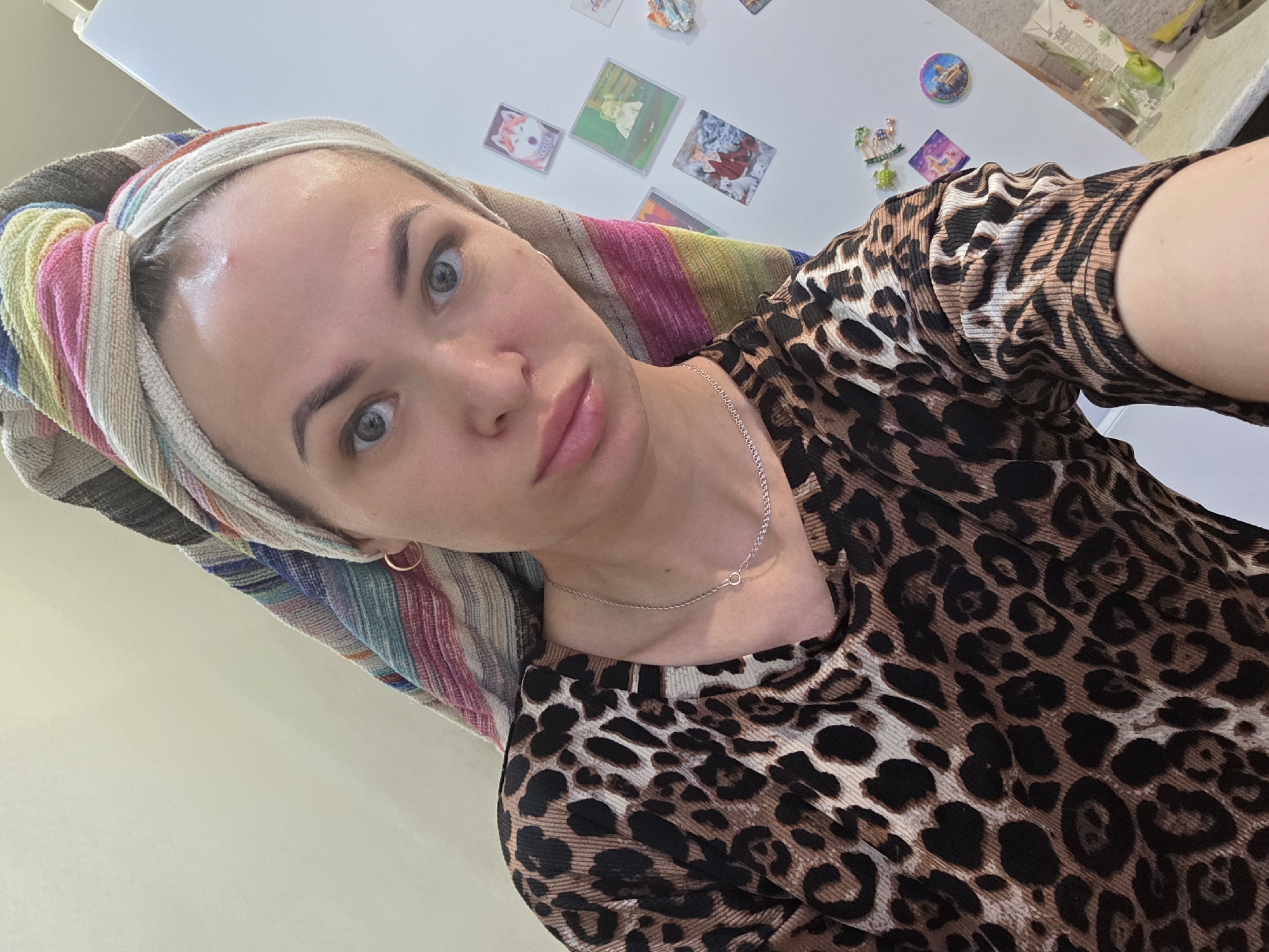 A woman is posing for a selfie in what appears to be an indoor setting, likely a home. She has a striped towel wrapped around her head. She is wearing a brown leopard print shirt and a silver necklace. Refrigerator magnets are visible in the background. There is no information suggesting the location is in the city of Chaikovsky, Russia.