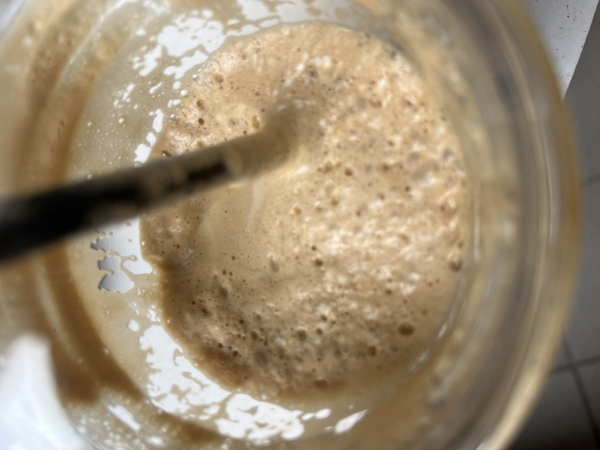 The image shows a close-up, top-down view of a foamy, light brown beverage in a clear glass. A dark straw or stirring stick is submerged in the center of the drink, creating ripples and disturbing the foam. The beverage appears to be a coffee-based drink, possibly a latte or cappuccino, with a significant amount of froth on the surface. The lighting suggests it might be daytime, with light reflecting off the glass and the surface of the drink. There are no people or discernible elements of the environment visible that would allow for identification of a specific location in Sofia, Bulgaria.