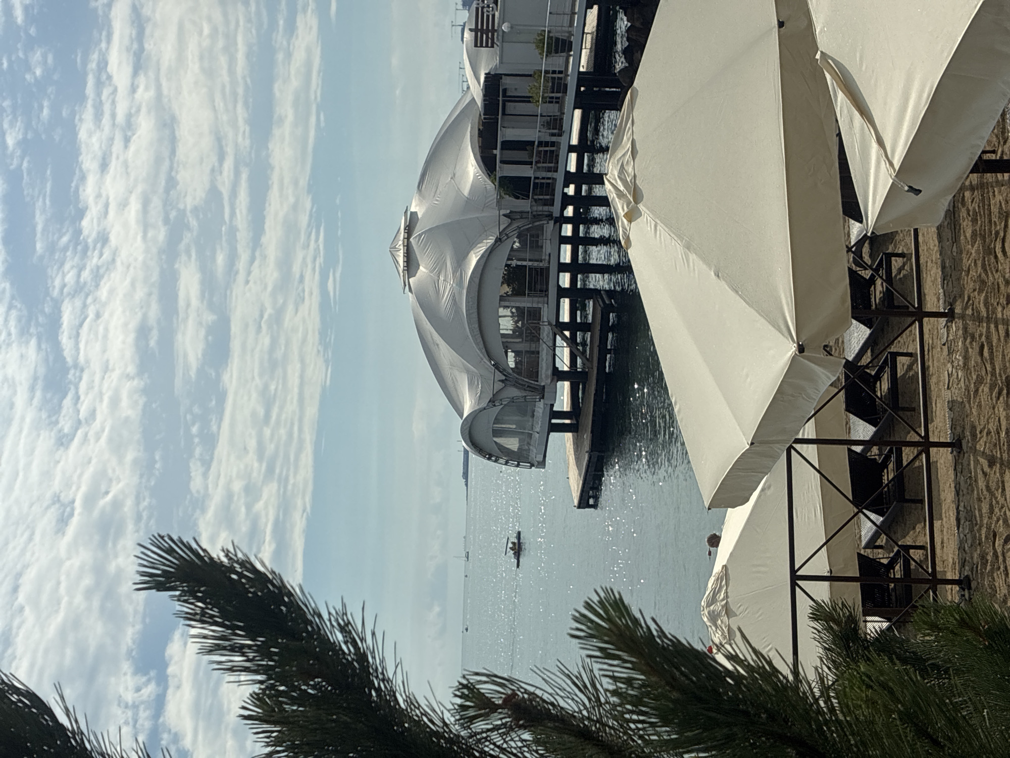 The image depicts a waterfront scene in Gelendzhik, Russia. In the foreground, several beach umbrellas and lounge chairs are set up on a sandy beach. Behind them, the sparkling water of the sea is visible, with a few boats in the distance. A distinctive white domed structure on stilts extends over the water, suggesting a restaurant or event space. The sky is partly cloudy, with a bright, sunny atmosphere. The presence of umbrellas and lounge chairs indicates a recreational area, likely a beach resort or public beach. The architectural style of the domed structure and the overall ambiance suggest a coastal tourist destination.