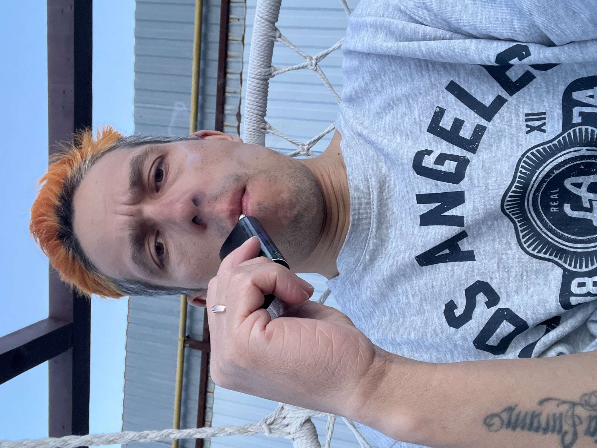 A fair-skinned man with dyed orange and gray hair is vaping. The man is wearing a gray shirt with "Los Angeles" printed on it. He is sitting in a rope chair outdoors. The background includes a building exterior with corrugated metal siding and a blue sky. The location is outdoors, and the city is stated to be Chaikovsky, Russia.