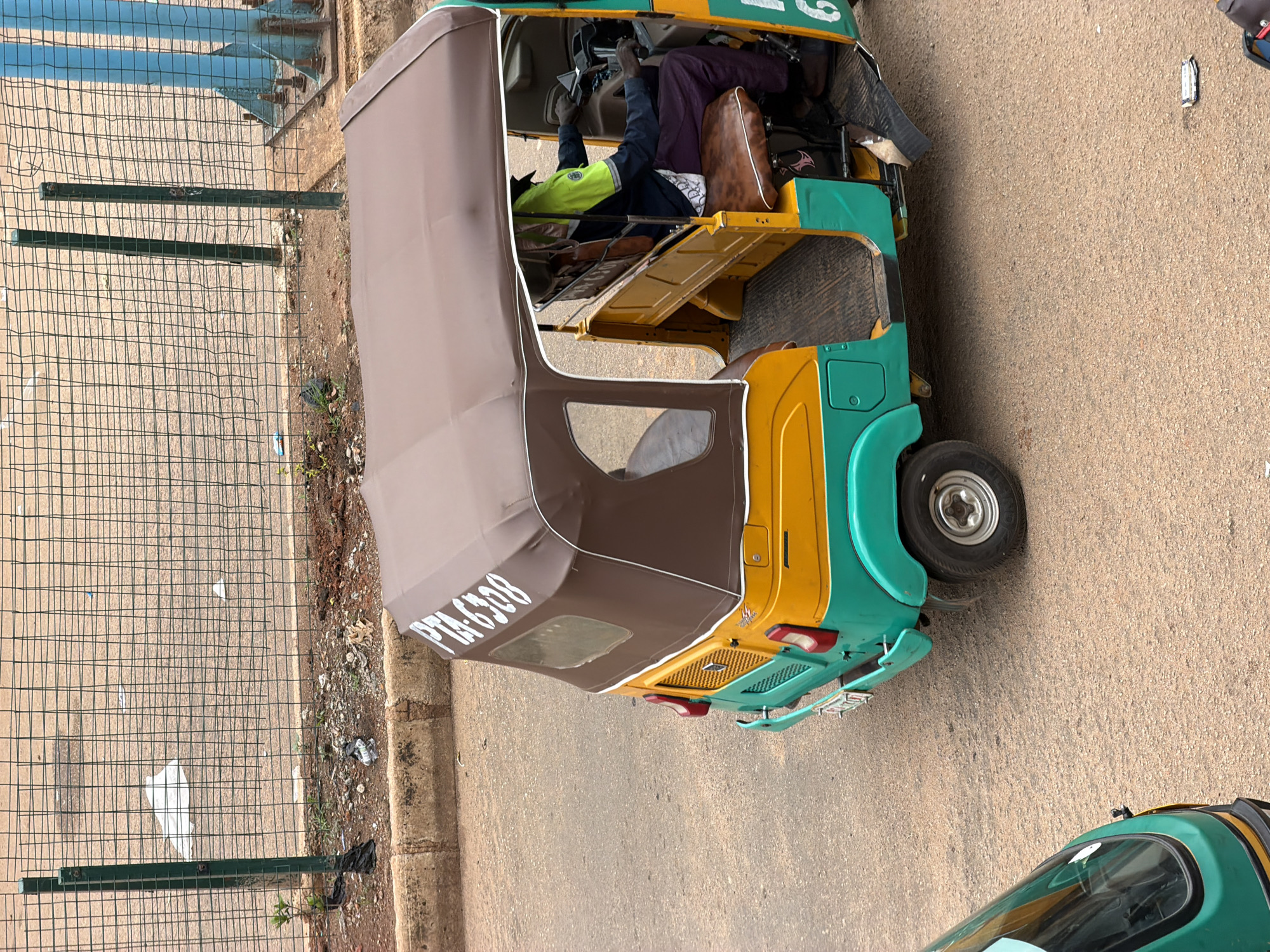 An elevated, eye-level medium shot captures a vibrant three-wheeled auto-rickshaw, commonly known as an "okada" or "Keke Napep," on a dusty road in Bukuru, Nigeria. The vehicle is predominantly teal green and bright yellow, with a brown canvas canopy. White lettering on the canopy reads "PTA-6308." A person, whose skin tone suggests they are of African descent, is seated in the driver's position, wearing a bright lime green, dark blue, and black jacket, and dark purple trousers. Their hands are on the steering wheel, and they appear to be operating the vehicle. The interior of the auto-rickshaw features wooden plank flooring and yellow-upholstered seating. A second, partially visible auto-rickshaw, primarily teal green, is positioned in the foreground, to the left of the main subject. In the background, a metal chain-link fence separates the road from a dirt area with sparse vegetation and some scattered litter. Beyond the fence, a road or walkway is visible, with a blue metal support structure and more fencing in the distance. The overall environment suggests a typical urban or peri-urban setting in Nigeria.