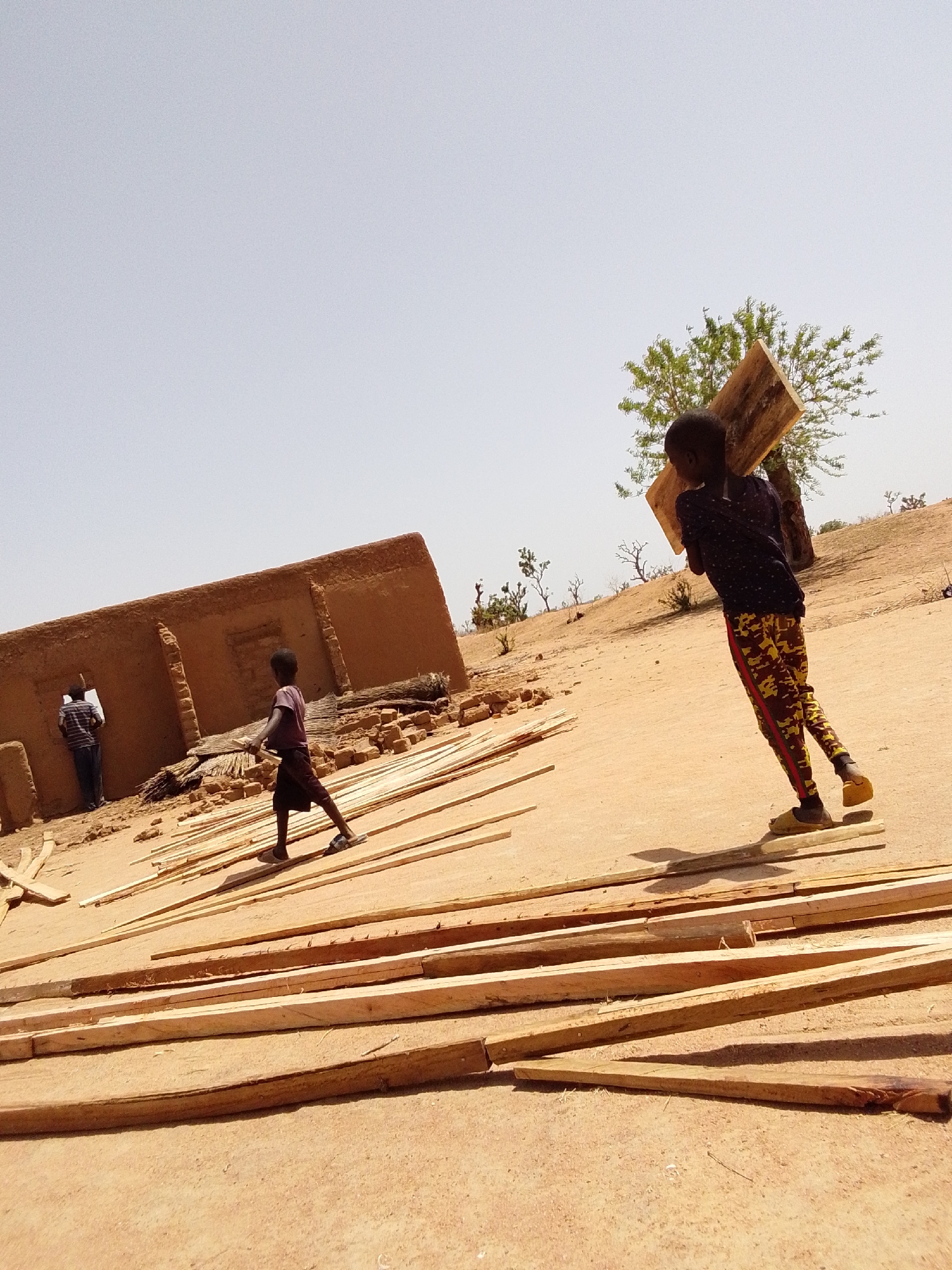 The image depicts a construction scene in what appears to be a rural area, likely in Mulke Barikin Sayejo, Nigeria. In the background stands a partially constructed or dilapidated mud building. In the foreground, multiple wooden planks are scattered on the sandy ground. A young individual, dressed in a dark patterned shirt and patterned pants, is carrying a large wooden board on their shoulder. Another child is walking towards the building. An adult stands beside the building, their activities unidentifiable. A sparse tree and sparse vegetation are visible in the backdrop under a clear, bright sky.
