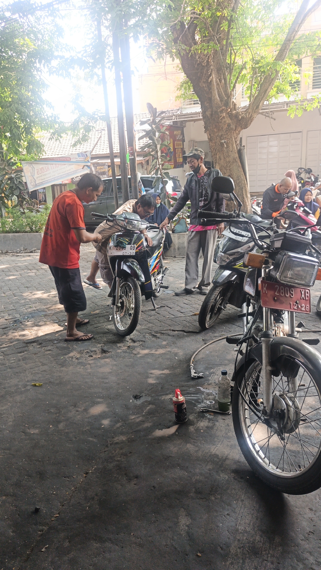 In Mlati Kidul, Indonesia, a group of individuals are gathered around motorcycles on a paved, somewhat unkempt surface. The scene appears to be an informal outdoor workshop or repair area.

In the foreground, a motorcycle with license plate "K 2809 XB" is partially visible, showing its front wheel and fender. To the left, a man in an orange t-shirt and black shorts is bent over another motorcycle, seemingly working on it. This motorcycle has a license plate that reads "K 2065 DB" with "07.30" below it.

A man wearing a dark jacket with a patterned shirt underneath, and a camouflage cap, stands behind the orange-shirted man, observing the work. Another person, partially obscured, appears to be a woman wearing glasses and a blue headscarf, leaning towards the motorcycle being worked on.

Further back, a man in an orange jacket and grey pants stands near a light-colored motorcycle. To the right, a person in an orange shirt with a dark head covering is seated on a motorcycle. Several other motorcycles are visible in the background, along with some buildings and trees. The ground is marked with oil stains and debris, suggesting it's a frequently used area for motorcycle maintenance. A can of what appears to be cleaning spray and a bottle of liquid are on the ground near the motorcycles. The overall impression is a bustling, everyday scene of motorcycle repair.