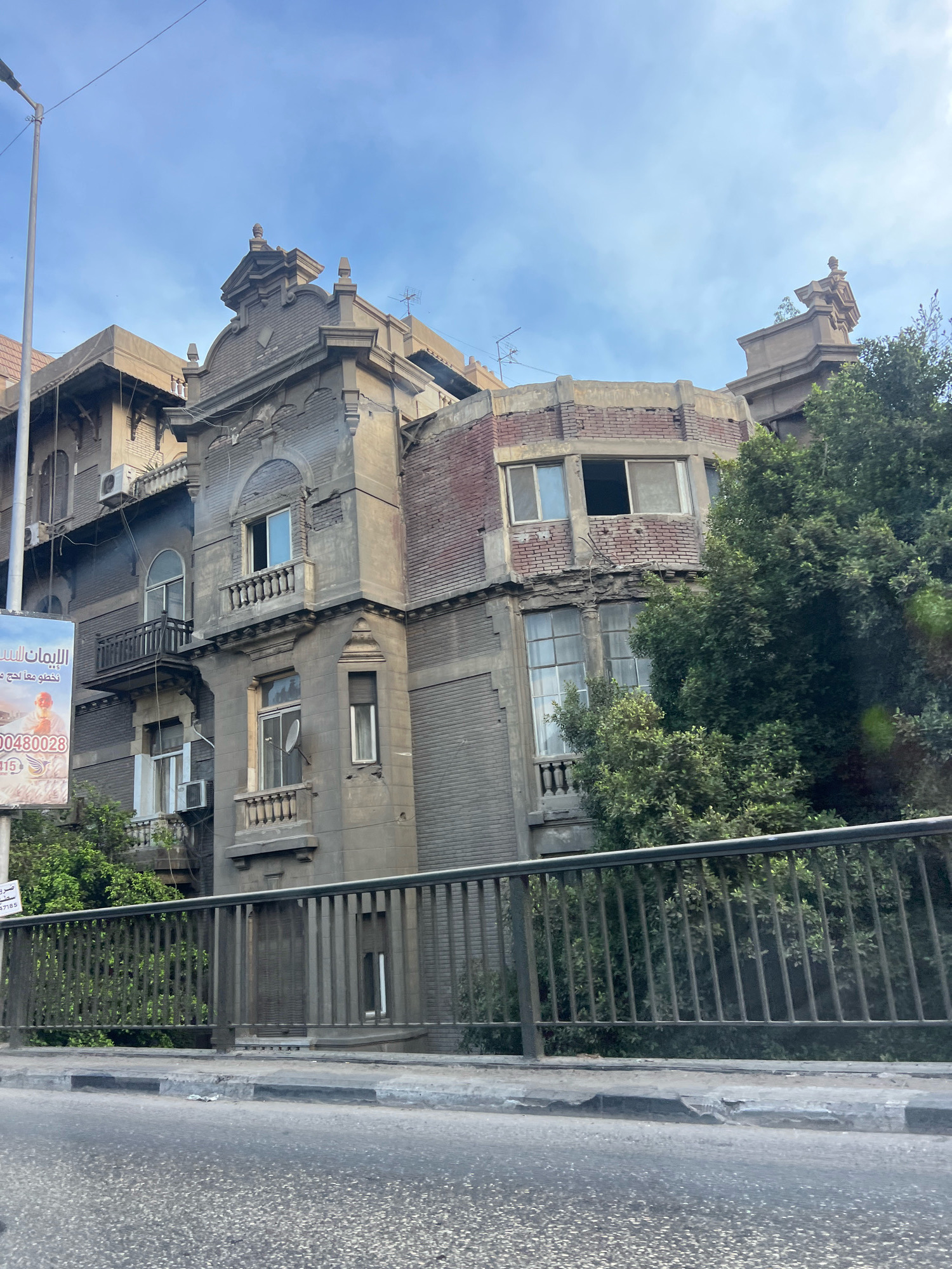 The photograph, taken in Unknown, Egypt, captures a historic building with ornate architecture, partially obscured by lush green trees and a metal railing in the foreground. The building exhibits signs of age, with weathered stone facades and visible brickwork on a curved section. Multiple windows, some with balconies, punctuate the structure. An advertisement banner is visible on the left side of the building. The sky above is partly cloudy, with a clear blue expanse visible. The foreground shows a street with a curb and a road surface, indicating an urban setting.