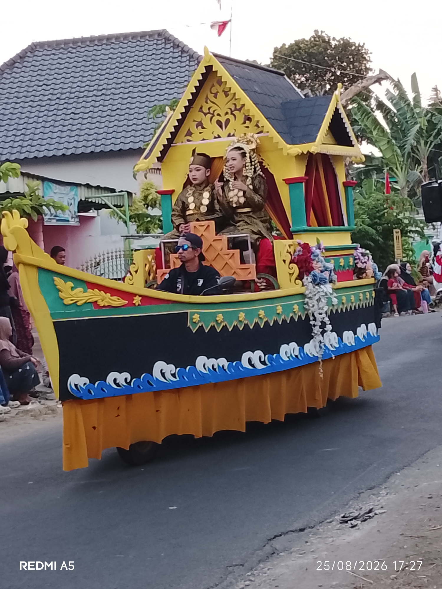 This image captures a festive parade float, designed to resemble a boat, moving along a street in Semanding, Indonesia. The float is elaborately decorated with a yellow, house-like structure on top, adorned with intricate yellow cutouts and red and green curtains. Two individuals, a male and a female dressed in traditional attire, are seated within this structure. The male wears a dark green outfit with gold embellishments and a hat, while the female is in a similar outfit, accessorized with elaborate jewelry and a headdress.
Below the seating area, the float is shaped like a boat's hull, painted black with a red and green trim along the top edge. Yellow decorative elements and floral motifs are visible on the green trim. A distinct wavy blue and white pattern adorns the lower part of the hull, with a vibrant orange skirt-like fabric draping the base, partially concealing the wheels.
A man in a black shirt, black baseball cap, and sunglasses is seated at the front of the float, appearing to be operating or guiding it. The scene is set against a backdrop of residential buildings with grey tiled roofs and lush green trees, including banana trees, indicating a tropical environment. Several spectators, including women in headscarves and children, are visible on the sides of the street, observing the parade. The overall atmosphere suggests a cultural celebration or local event. The timestamp "25/08/2026 17:27" and "REDMI A5" are watermarked on the image.