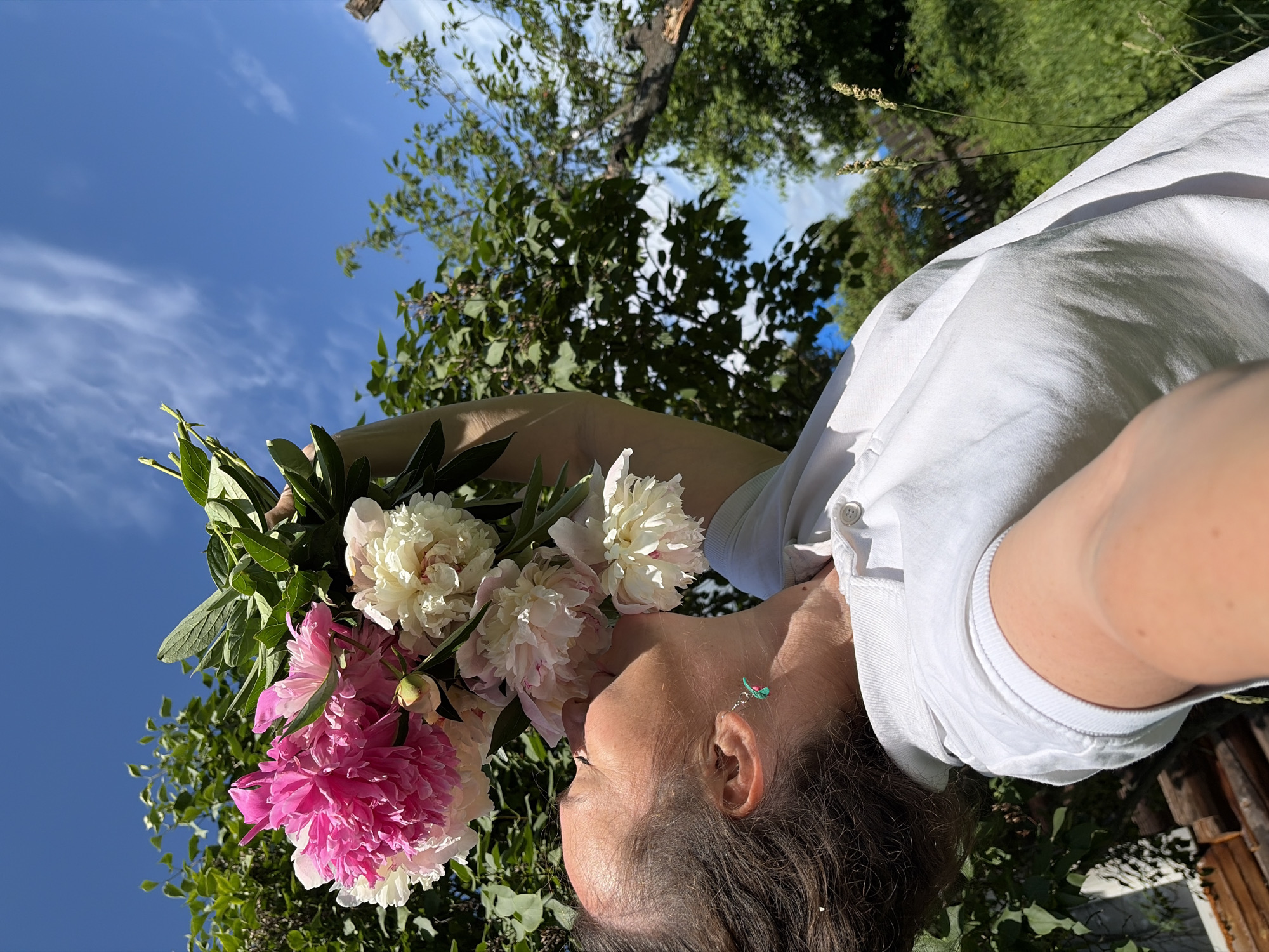 A close-up, overhead shot shows a person wearing a white shirt and holding a bouquet of pink and white peonies. The person's head is tilted downwards, and they are looking towards the flowers. A small, colorful earring is visible on their ear. The background consists of a bright blue sky with scattered clouds and lush green trees. The scene appears to be set outdoors on a sunny day, likely in a garden or natural setting. The lighting suggests it is daytime. The image is captured from a high angle, looking down on the subject and the flowers. No text is visible in the image.