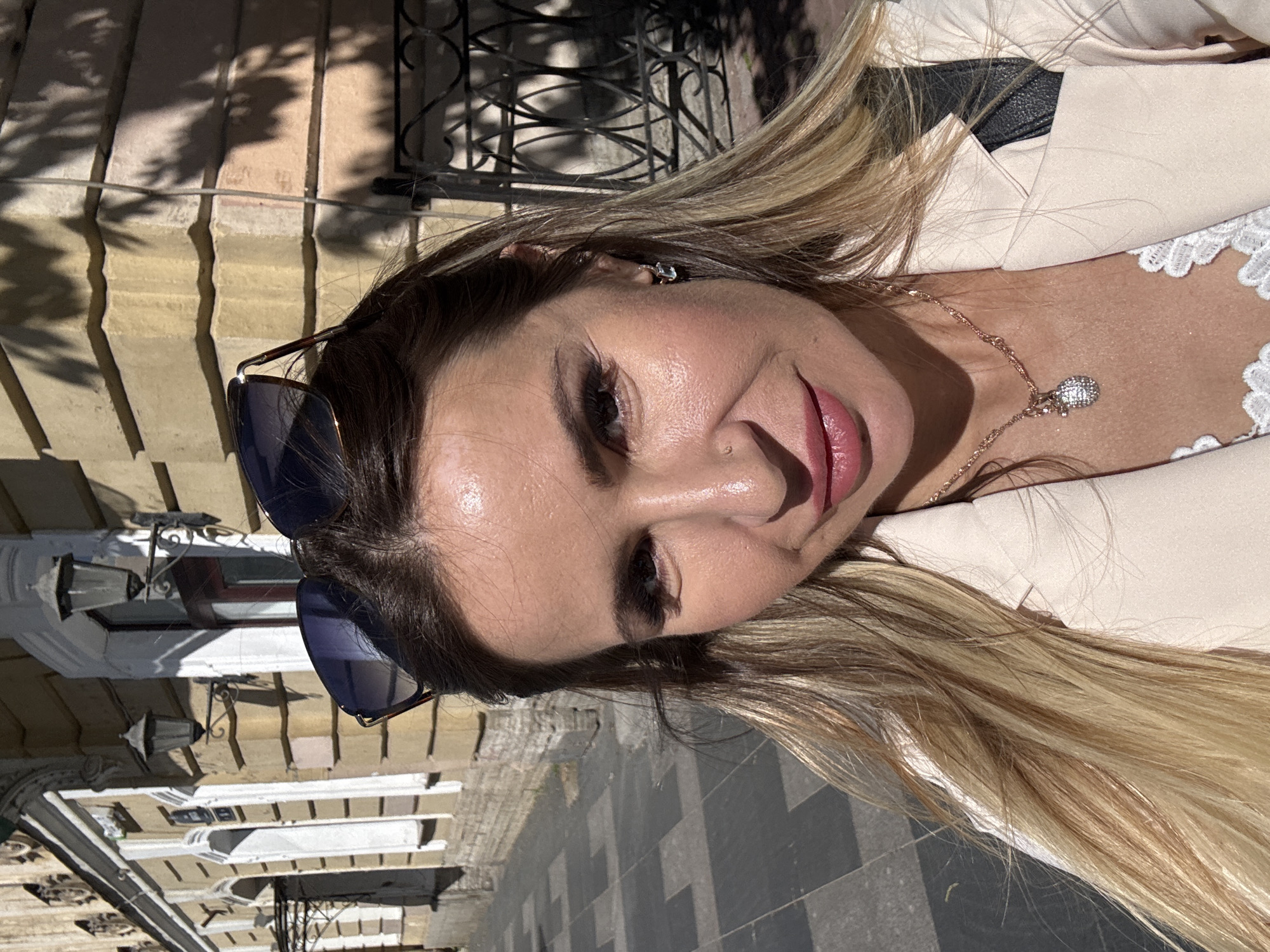 A woman stands on a sidewalk in Saint Petersburg, Russia, posing for a selfie in front of a building. The building's facade is yellow with white architectural details and ornate black street lamps. The sidewalk is paved with dark and light square tiles. The woman has long, blonde hair and is wearing a light-colored blazer over a white lace top. She is also wearing sunglasses perched on her head and a necklace with a pendant. The sun casts shadows from tree leaves on the building.