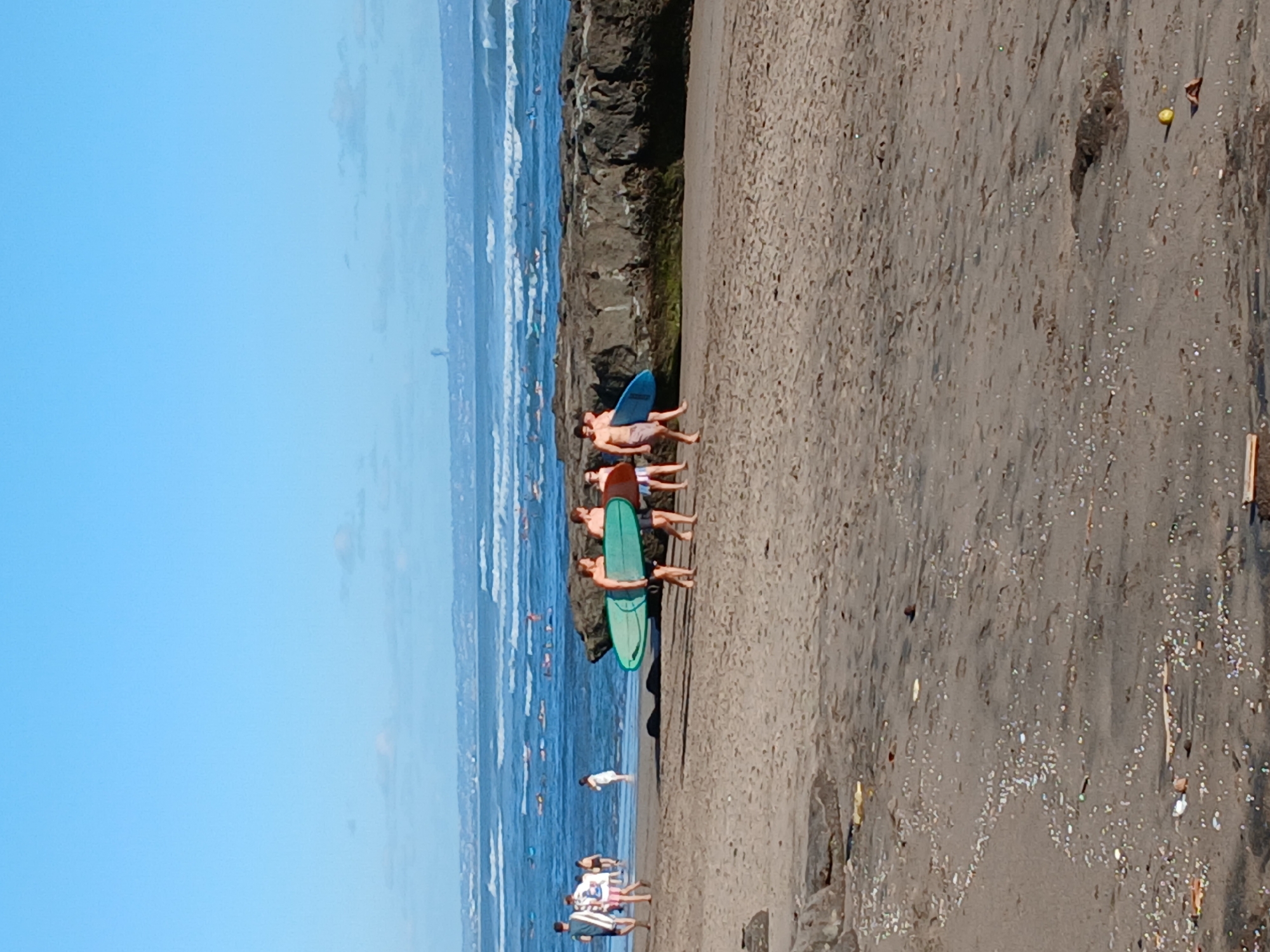 Four individuals are walking on a sandy beach in North Kuta, Indonesia. Two men are carrying surfboards. The man on the left is holding a light green surfboard and the man to his right is holding a darker teal surfboard. Two other individuals, a man and a woman, are walking behind them carrying surfboards. The ocean is visible in the background with several surfers in the water. The sky is clear and blue. The beach is composed of sand with some scattered rocks and debris.