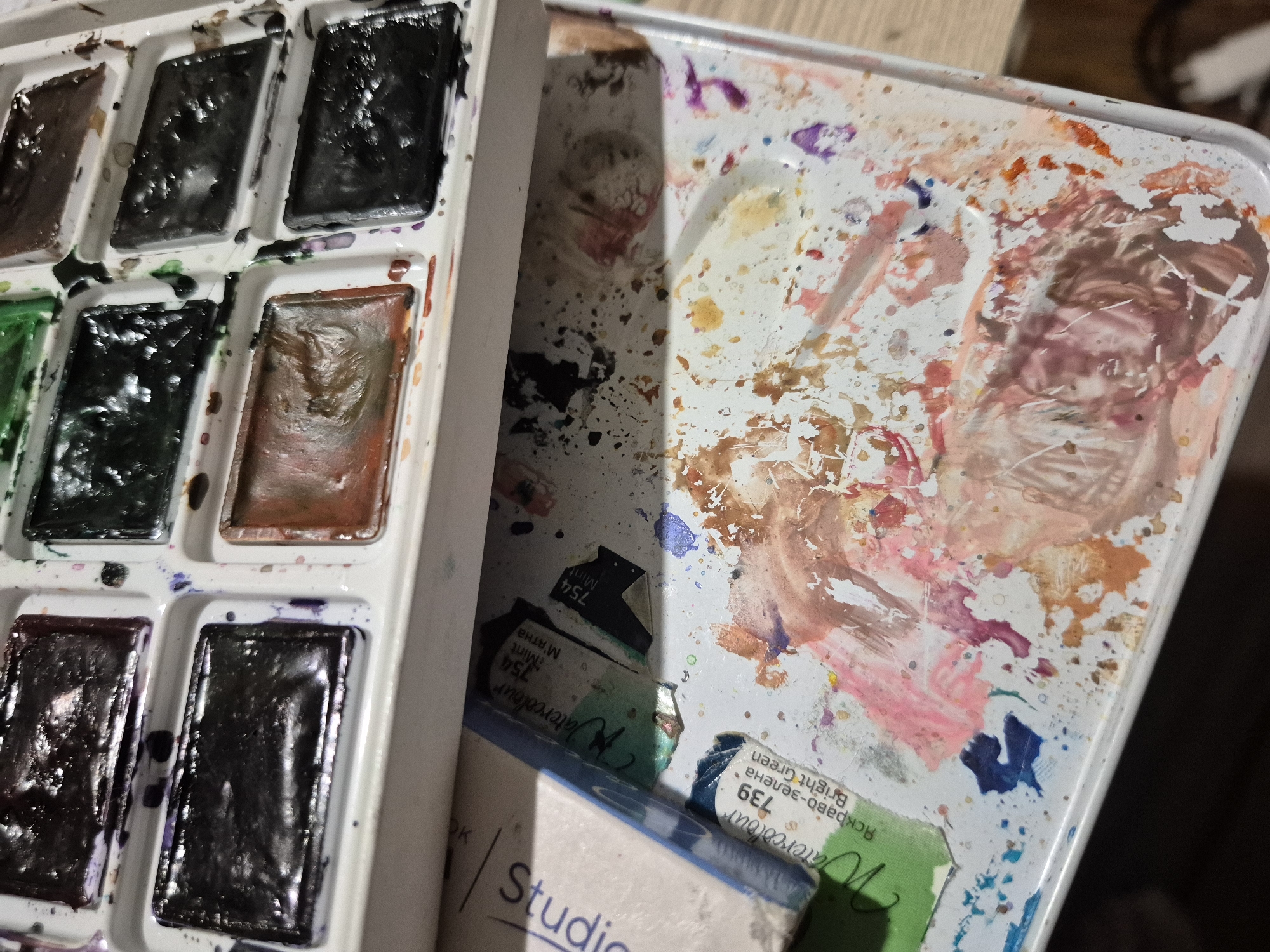The image displays a close-up, eye-level, indoor shot of a used watercolor paint palette and its associated supplies. The palette itself is white and has multiple rectangular wells filled with various colors of dried watercolor paint, including dark purples, blacks, greens, and browns. Splatters and streaks of wet and dried paint are visible across the palette, indicating recent use. Below the palette, there is a white surface with further paint splatters and smears in shades of pink, orange, blue, and purple. Partially visible in the lower left corner are two small paint tubes. One tube is labeled "Watercolour 739 Яскрaво-зелена Bright Green," and the other, slightly out of focus, is labeled "Watercolour 754 Mint" with Cyrillic script. The overall impression is that of an artist's workspace, with materials in active use. The scene is devoid of any people or signs of life. The image does not provide any information that would allow for the localization of this scene in Popovo, Bulgaria, or any other specific city.