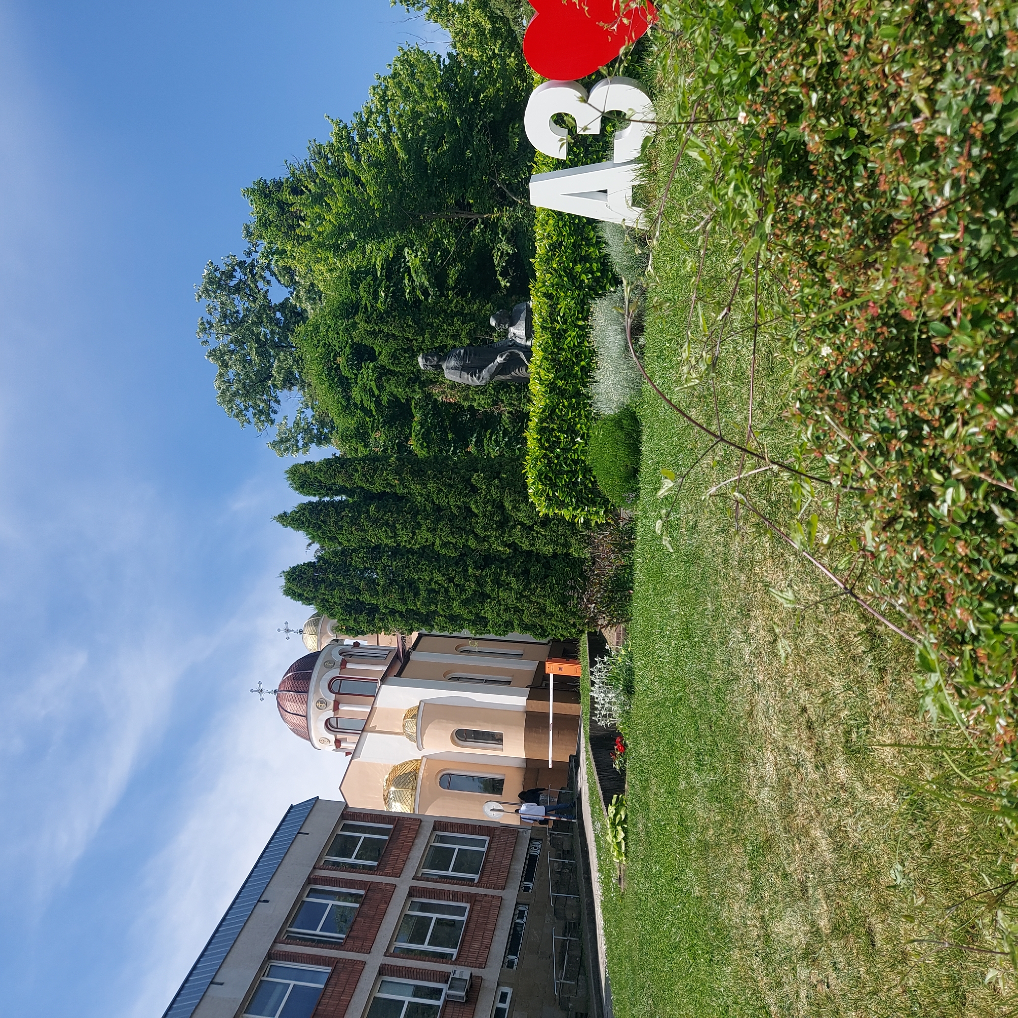 The image captures a sunny day in Veliko Tarnovo, Bulgaria. A red heart and white letters "A3" stand in a park near a statue and tall green trees. In the background, a church with a red dome and gold crosses stands next to a brick building with white-framed windows. People are near the church. The lawn in the foreground is green.