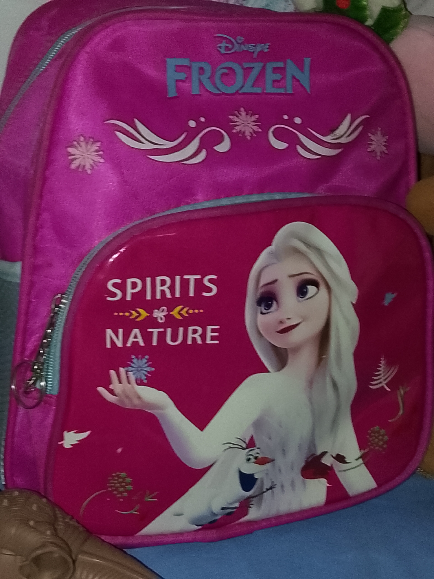 This media file displays a fuchsia-pink children's backpack, likely featuring characters and themes from Disney's "Frozen." The backpack has two main sections visible.
The larger, upper section of the backpack is pink/fuchsia and features the "Disney" logo in blue text above the word "FROZEN," also in blue, stylized block letters. Below the "FROZEN" text, there are white decorative swirls resembling ice or wind patterns, interspersed with several white snowflakes of varying sizes.
The smaller, front pocket of the backpack is also fuchsia-pink and prominently features Elsa from "Frozen 2" and Olaf the snowman. Elsa is depicted on the right side of this pocket, facing left with her head turned to look upwards and to the right, sporting a serene smile. She has long white hair and is wearing a white garment. Her right hand is extended, holding a light blue snowflake. Olaf is positioned near Elsa's lower torso, looking forward with a cheerful expression.
Text on this front pocket reads "SPIRITS" in white, followed by a line of decorative white and yellow arrow-like and leaf-like symbols, and then "NATURE" in white text. The design also includes various natural elements such as a small white bird or leaf, clusters of red berries or pine cones, abstract tree-like shapes, and a small blue butterfly or moth.
The backpack features a silver-colored zipper and pull tab on the front pocket. The material appears to be fabric. The backpack is resting on a blue surface, possibly a blanket or floor. Partial views of other objects, including a brown, textured object in the bottom left corner and some softer, possibly plush, items in the top right corner, suggest an indoor setting. There are no specific cues for time of day or weather, nor any direct connection to Davao City, Philippines, visible in the image.