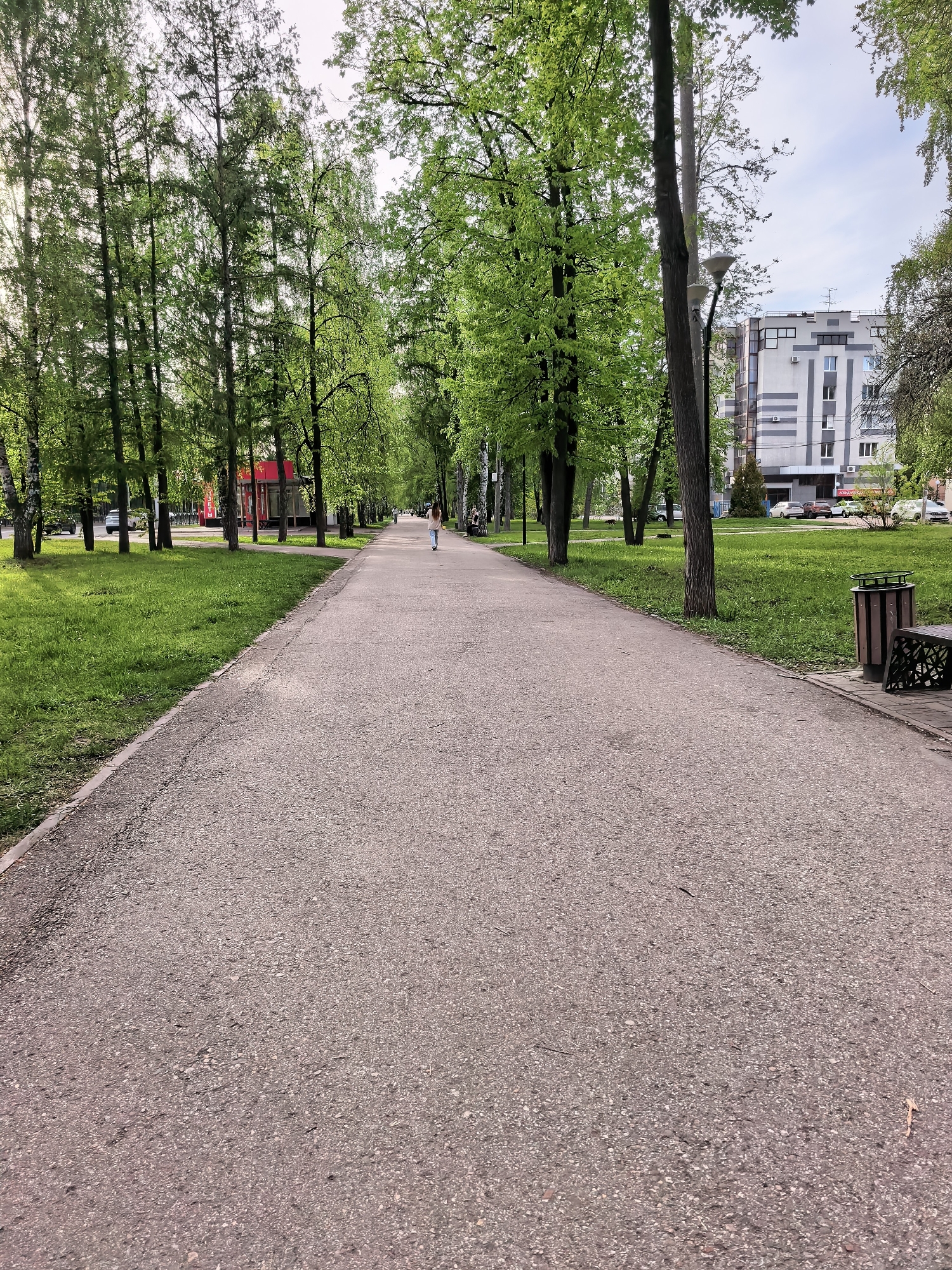 A medium-angle shot depicts an asphalt pathway stretching into the distance, flanked by green grass and trees on both sides. In the distance, a person is walking away. The path extends towards a multi-story building on the right. A bench with a trash receptacle is visible on the right side of the path. The location is Sterlitamak, Russia.