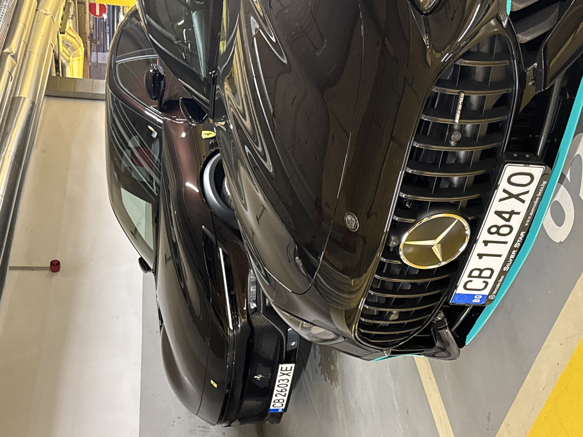 The image captures two parked cars, likely in a parking garage, with a focus on their front ends. The vehicle on the right is a Mercedes-Benz AMG, distinguished by its prominent Mercedes emblem on the grill and the Bulgarian license plate "CB 1184 XO." Next to it, on the left, is a Ferrari, identifiable by its unique design and Bulgarian license plate "CB 2603 XE." The cars are parked close together. The environment suggests an indoor parking facility, with visible pipes and lines along the wall, typical of such structures. The presence of Bulgarian license plates ("CB") indicates the location is likely Sofia, Bulgaria.