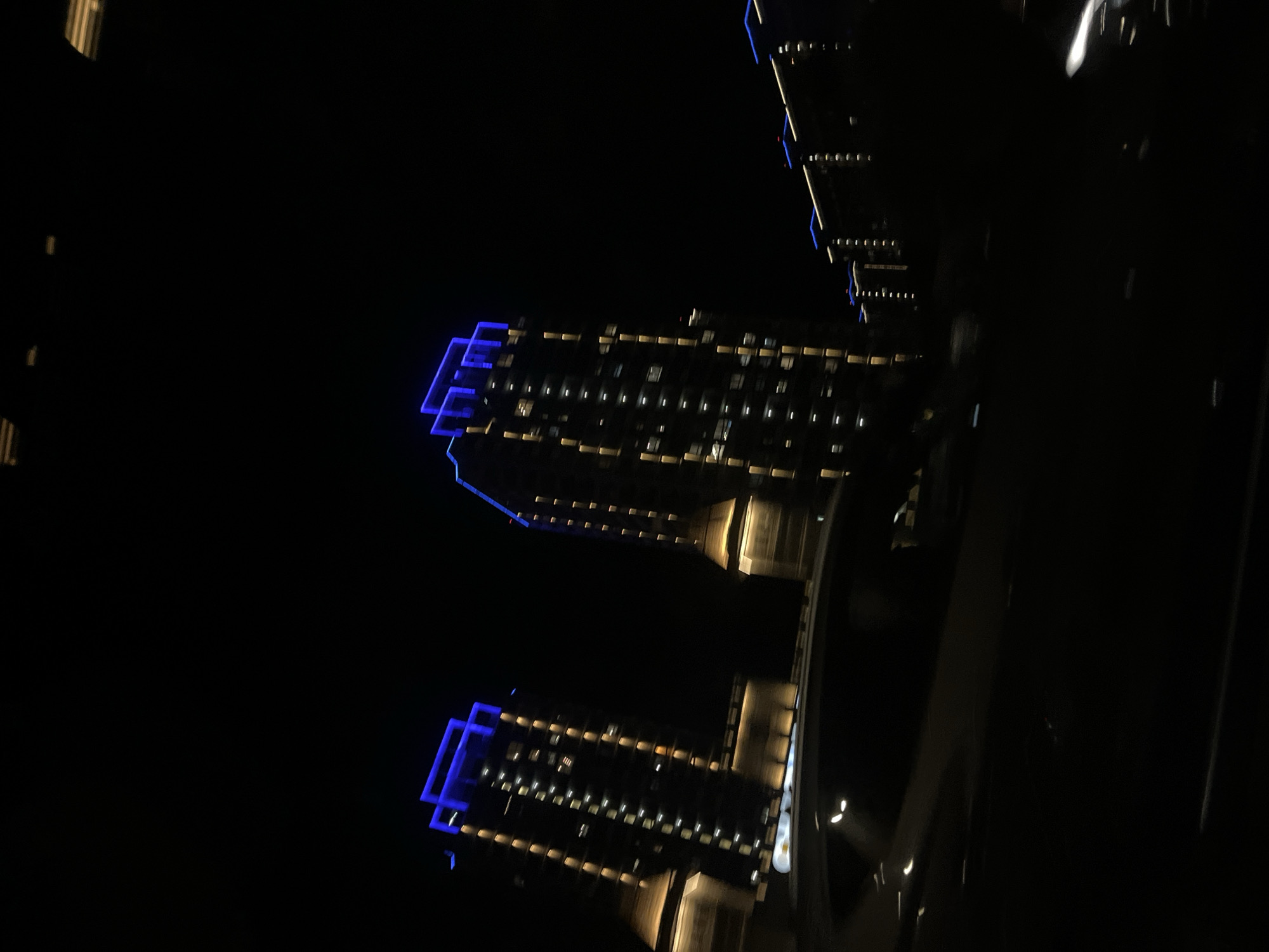 The media file displays a nighttime scene featuring two modern, illuminated buildings, likely located in Krasnodar, Russia. The image is rotated counter-clockwise, showing the structures angled upwards from the bottom right.
The primary subjects are the two tall buildings, characterized by their contemporary architecture. They are set against a completely dark sky, indicating it is late at night. The buildings are brightly lit, with a striking contrast between cool blue and warm yellow-orange light. Blue LED-like strips highlight the upper sections and architectural edges of both buildings, creating geometric patterns. Numerous windows on each building emit warm yellow or orange light, suggesting occupied residences or offices. The lower sections of the buildings also feature warm-toned lighting.
No specific human activity or event is visible. The scene is static, capturing the aesthetic of urban night-time illumination. The weather appears clear, with no clouds or atmospheric obscurities visible against the dark background. There is no discernible text within the image. The overall impression is one of modern city architecture illuminated for the evening.