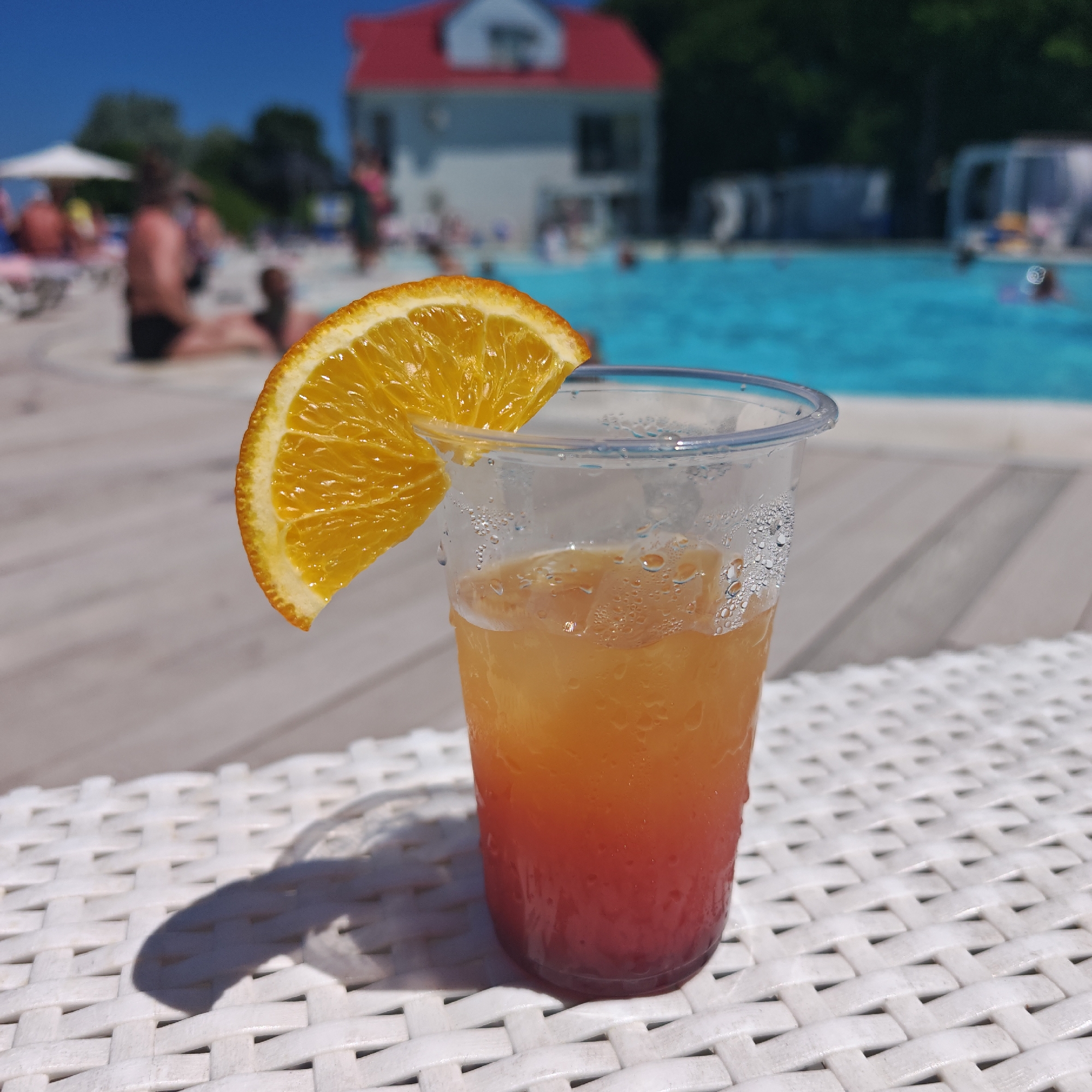An outdoor scene with a drink in the foreground. The drink is in a clear plastic cup and has a layered orange and red color, garnished with a slice of orange. The cup has condensation on the outside. It rests on a white woven surface. In the background, a swimming pool with people is visible, along with some buildings and trees under a clear blue sky. The location is identified as городское поселение Щелкино, Ukraine.