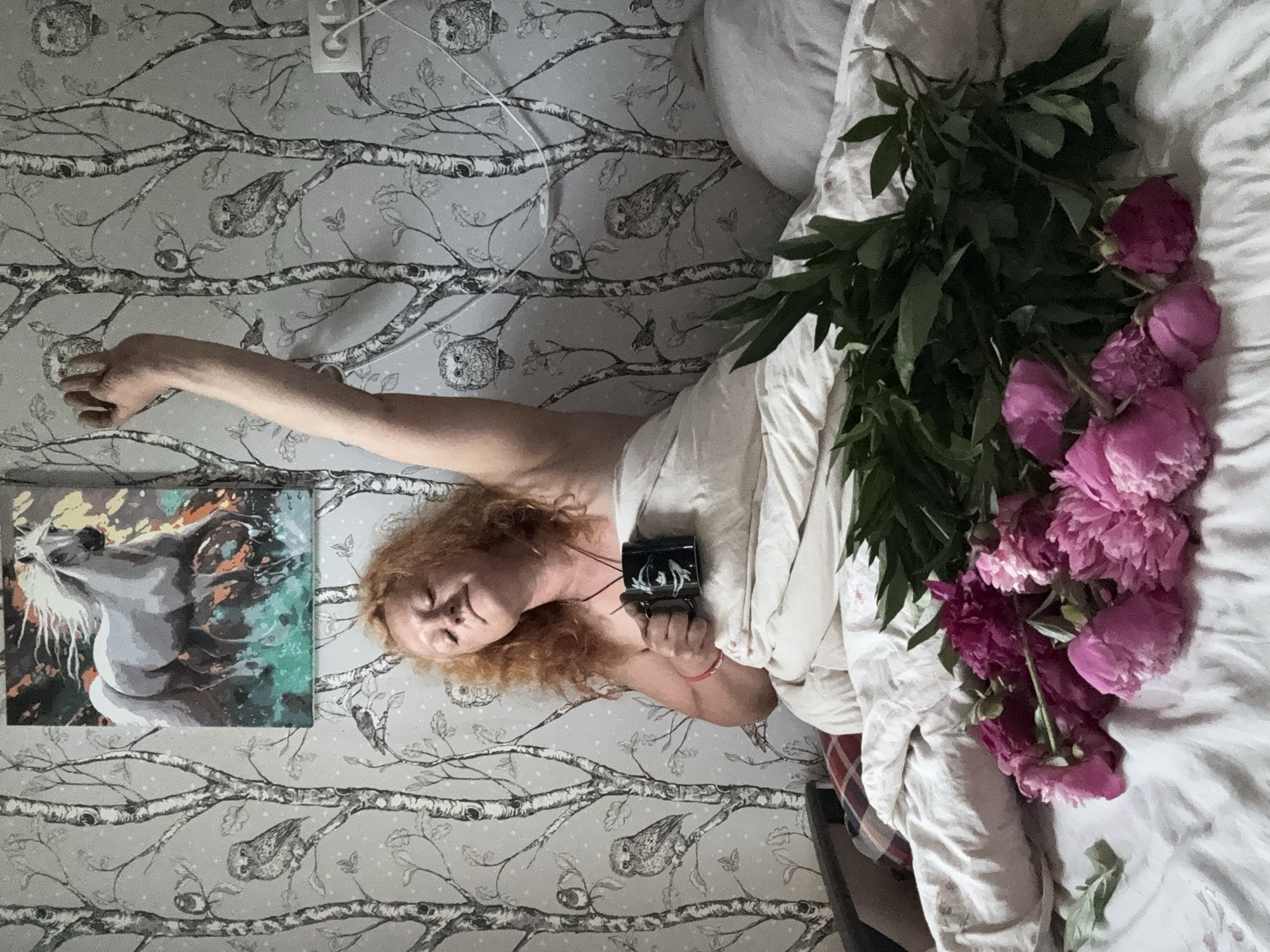 A woman with red hair, wrapped in a white duvet, sits on a bed with her right arm raised and her eyes closed. She holds a black mug in her left hand. In the foreground, a bouquet of pink and magenta peonies lies on the white bedsheets. The wall behind her is decorated with wallpaper featuring a pattern of birch trees and owls. A framed painting of a white horse is hung on the wall above and to the left of the woman. The scene appears to be indoors, in a bedroom. The location is a bedroom in the city of Chaikovsky, Russia.