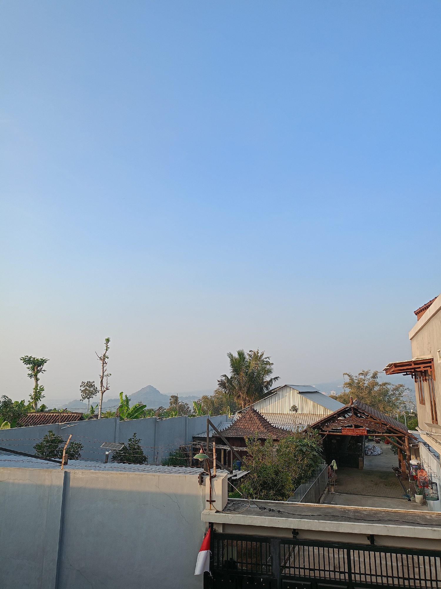 A wide-angle, eye-level shot depicts a residential area in Batu, Indonesia, under a clear, pale blue sky. The foreground features a gray concrete wall with a black metal fence on top. To the right, a sliver of a modern building's exterior, painted in a light beige with dark brown wooden accents and an awning, is visible. In the mid-ground, a lower gray concrete wall runs horizontally, topped with barbed wire and a small solar panel. Beyond this wall, a cluster of single-story buildings with traditional Indonesian tiled roofs and a more modern metal-roofed structure are nestled among lush green trees and banana plants. In the distance, rolling hills and a mountain peak are faintly visible through a hazy atmosphere. The scene is devoid of people.