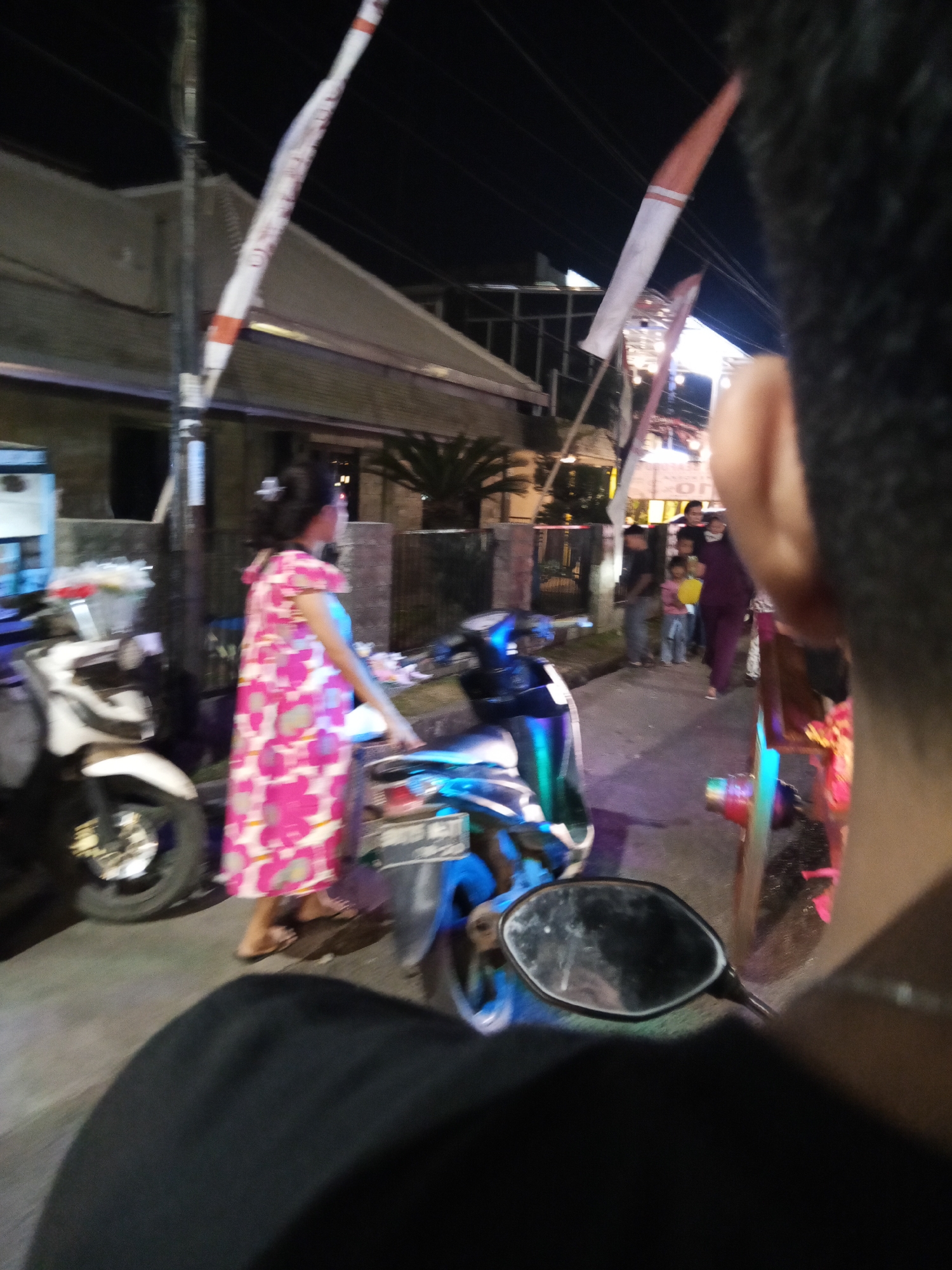 This is a photograph taken at night in the city of Lubuklinggau, Indonesia, depicting a street scene. The foreground is dominated by the shoulder and head of a person, suggesting the photo was taken from the perspective of someone riding on a motorcycle. A motorcycle mirror is visible in the lower right quadrant, reflecting a distorted image of the scene.
In the mid-ground, a woman in a vibrant pink and white floral dress is standing next to a white scooter. She is looking towards the right side of the frame. A blue scooter is parked in front of her. Further back, a group of people, including children, are visible on the sidewalk, some appearing to be interacting. Buildings with pitched roofs line the street, and there are lights illuminating the scene, typical of a commercial or residential area at night. Tall, narrow flags or banners, possibly for a local event or promotion, are prominently displayed on poles along the street. The overall lighting suggests artificial light sources from streetlights, buildings, and possibly neon signs.