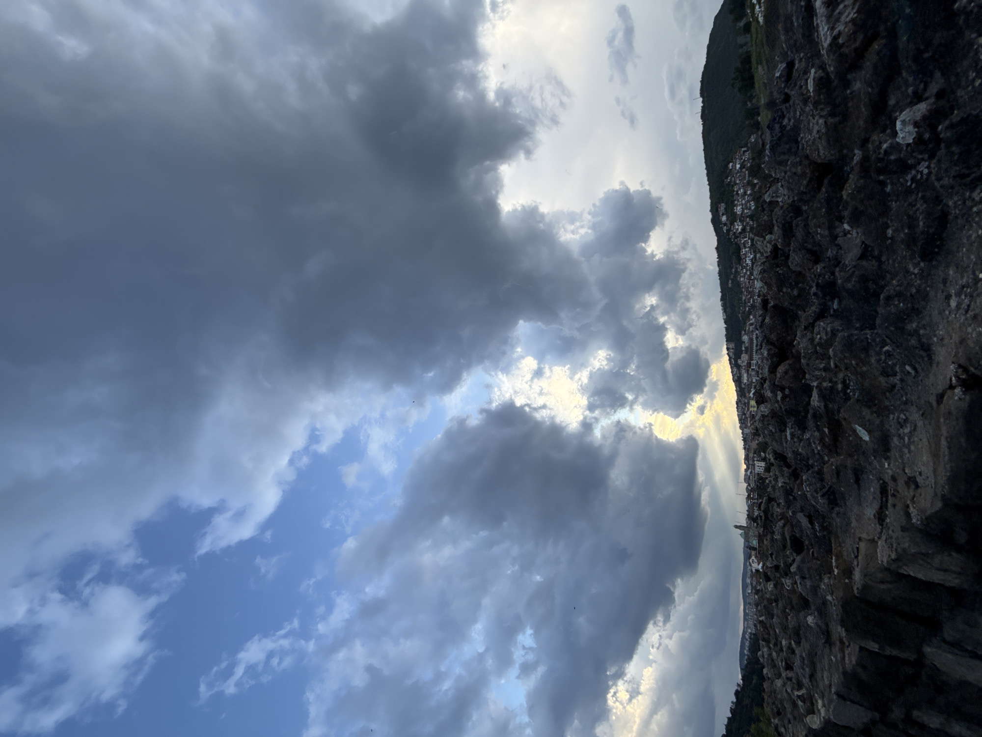 This photograph captures a dramatic sky filled with dark, gray clouds interspersed with patches of bright blue, suggesting an approaching storm. Rays of sunlight break through the cloud cover in the background.
The scene includes a portion of Veliko Tarnovo, Bulgaria, with buildings and trees visible on the hillside, partially obscured by the clouds. The foreground is dominated by a dark, rocky structure, possibly a wall or cliff, suggesting the photo was taken from a high vantage point.