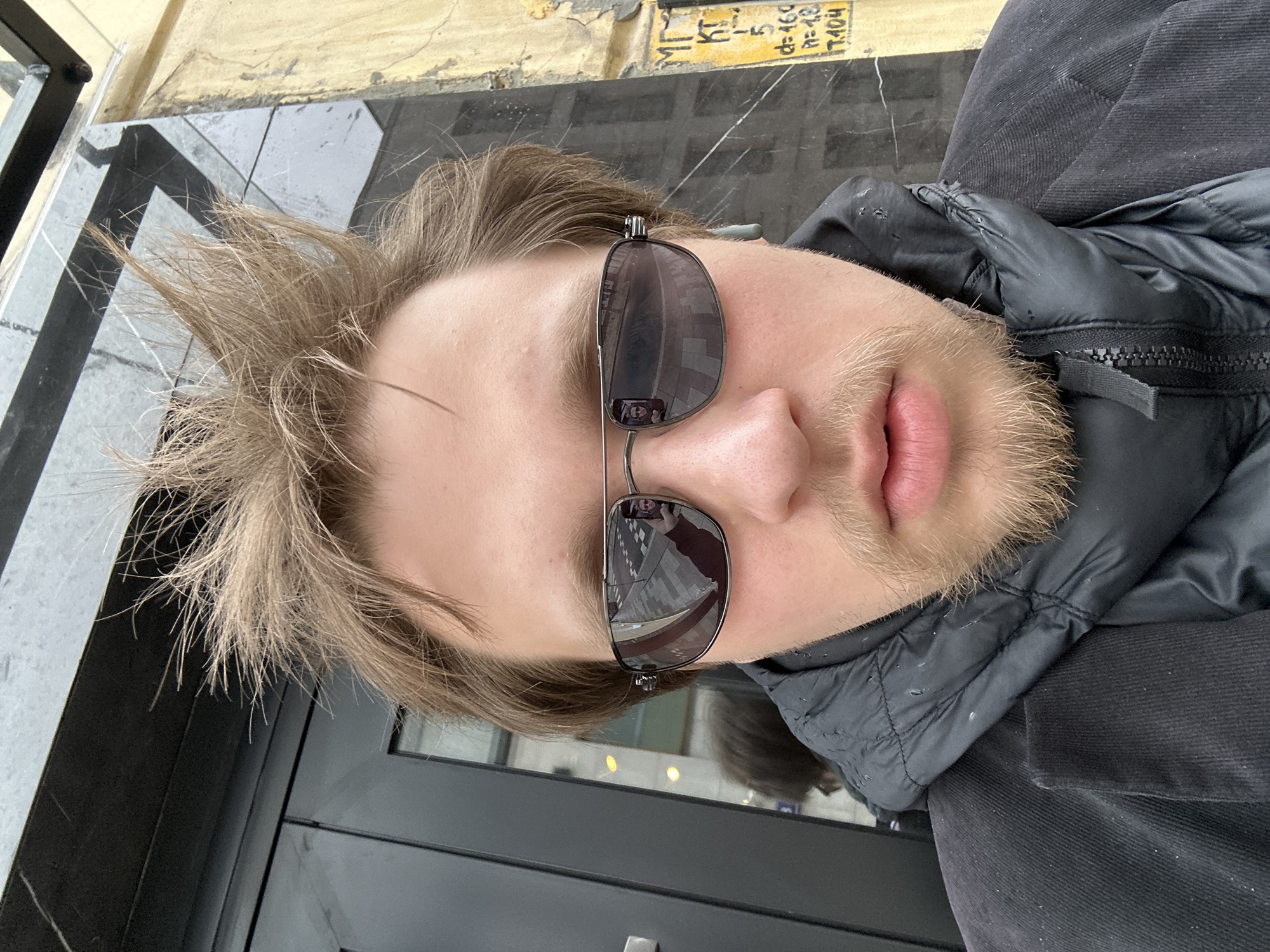 A close-up, vertically oriented shot depicts a person wearing sunglasses, with the nose positioned centrally in the frame. The person has light brown hair, a light beard, and light-colored skin. They are wearing a black, quilted jacket or vest, partly zipped up. The sunglasses reflect a city scene, visible in the lenses. The background consists of a wall with a dark, possibly metal, vertical element, and above it, a textured, pale-colored surface with what appears to be writing or markings. Based on the reflection, it seems to be a city in Unknown, Unknown.