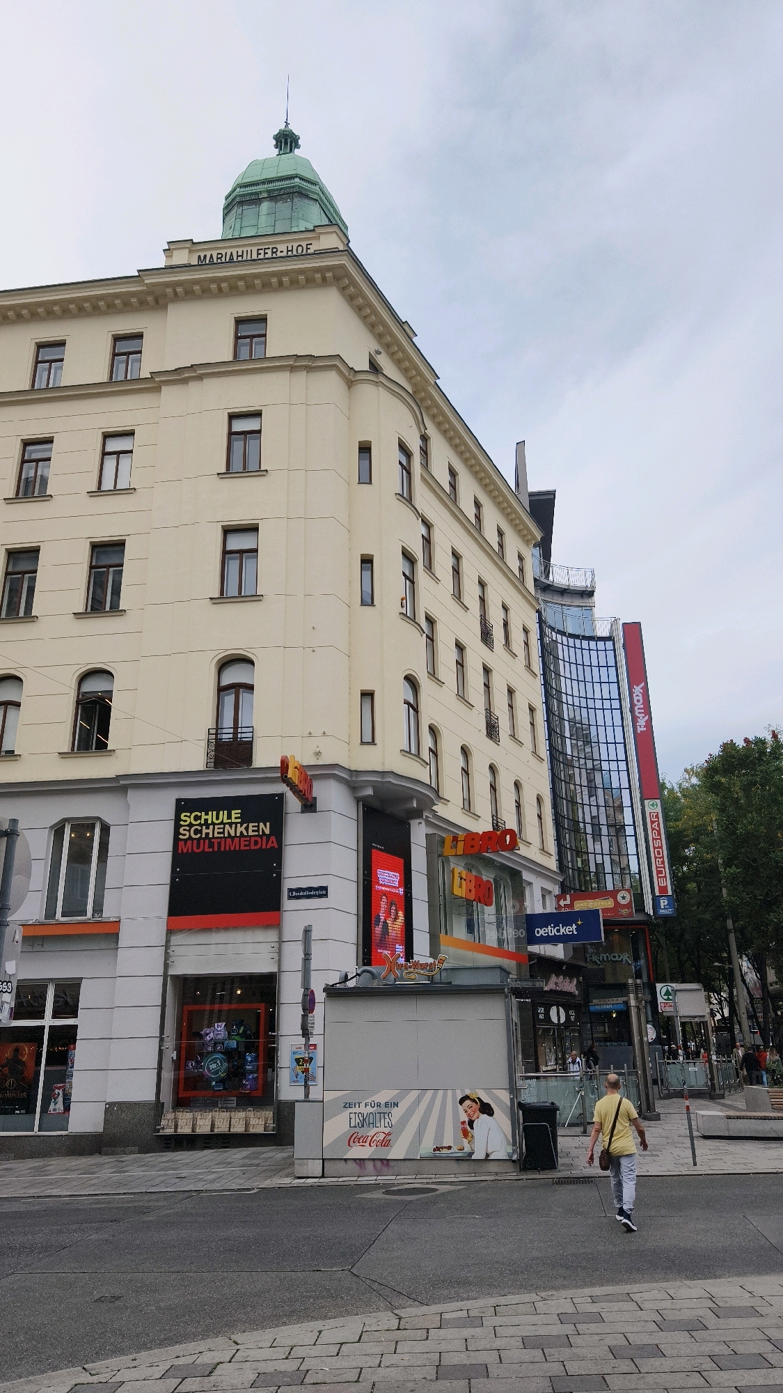 The image shows a street scene in Vienna, Austria, with a focus on the corner of a cream-colored building and its ground-level businesses. The building has multiple stories with evenly spaced windows. Atop the building, a green dome is visible, with text above it reading "MARIAHILFER-HOF."
On the ground floor of the building, there are several businesses. On the left, a sign reads "SCHULE SCHENKEN MULTIMEDIA." Next to it, a store called "LIBRO" is visible with red lettering. Another sign for "LIBRO" is present further down. Adjacent to these are signs for "oeticket," "TK MAX," and "EUROSPAR."
A man wearing a yellow shirt and grey pants is walking away from the camera on the right side of the street. A kiosk with a Coca-Cola advertisement featuring a woman is located in the foreground on the right. The street is paved with cobblestones and asphalt. The sky is overcast. The overall impression is a typical urban streetscape with commercial activity.