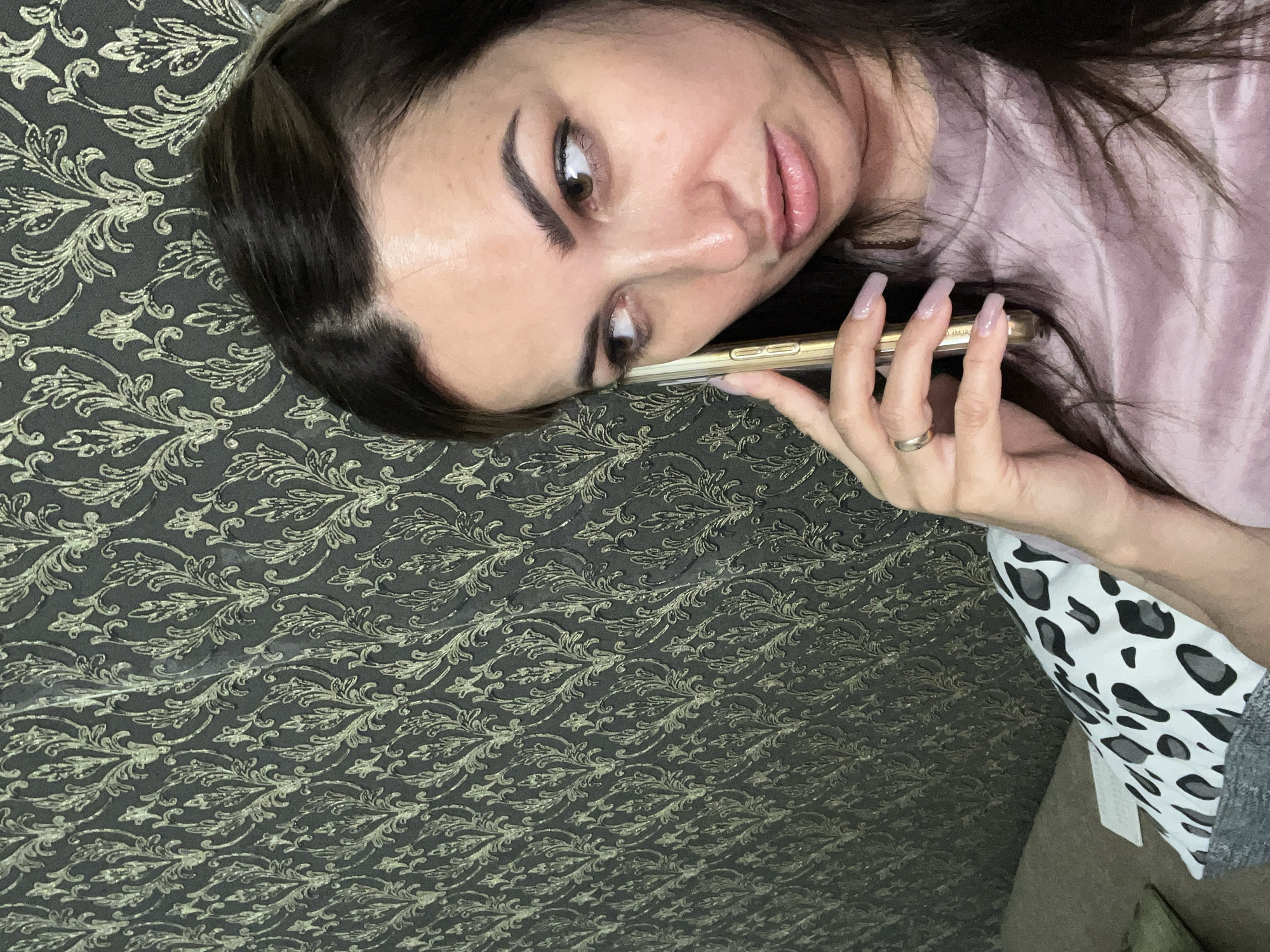 A woman with dark hair and brown eyes, wearing a pink top and a gold ring, holds a gold smartphone to her ear while looking to the left. She is positioned in the foreground, with a patterned wallpaper and a leopard-print pillow visible in the background.
