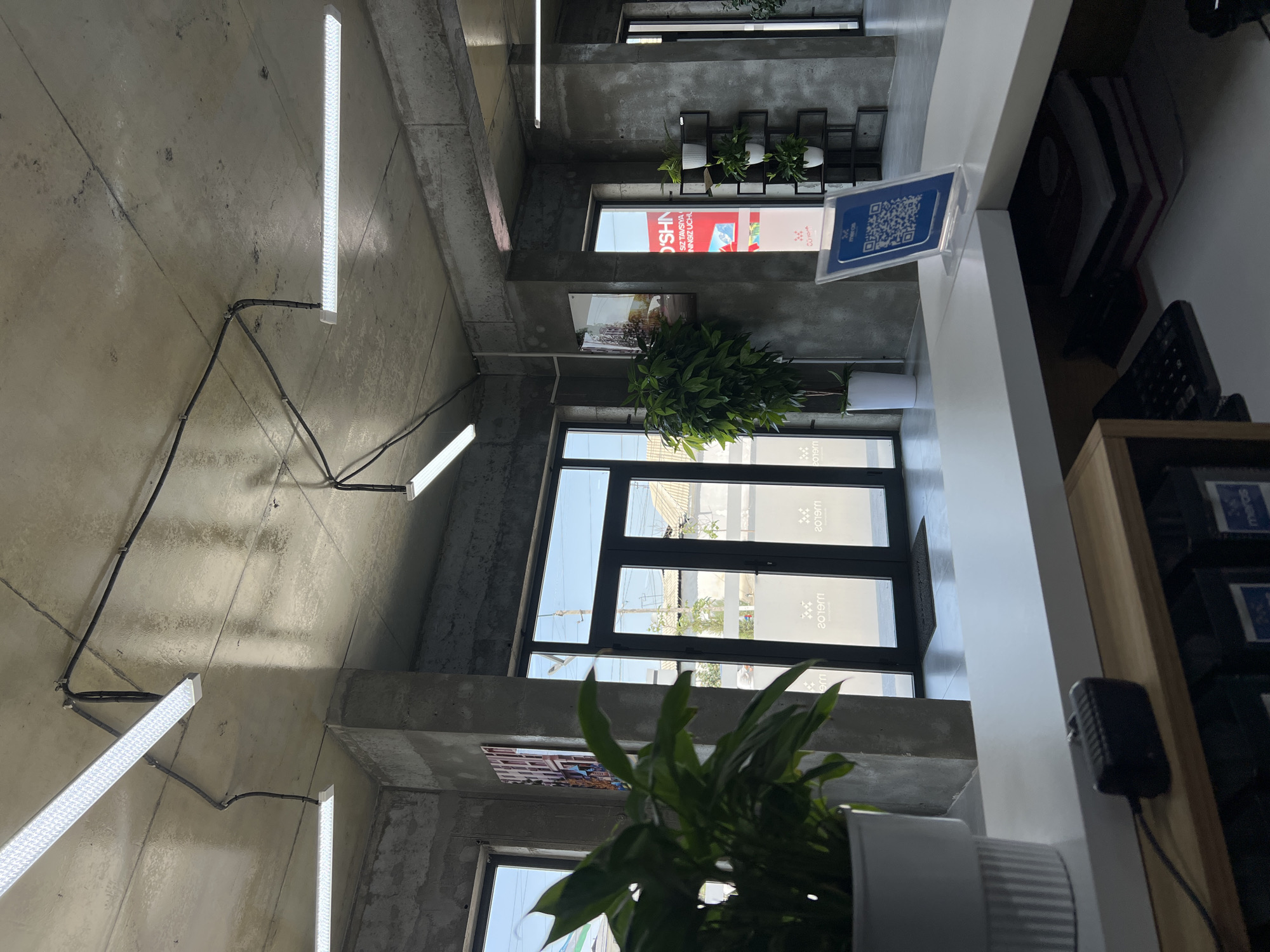 The image depicts the interior of a modern office or reception area in Shahrixon, Uzbekistan. The space features exposed concrete walls and ceiling, characteristic of industrial or minimalist design. Several long, rectangular fluorescent light fixtures are mounted on the ceiling, with visible electrical conduits running across.
In the foreground, a white reception desk is partially visible. On the desk, a small black electronic device, possibly a scanner or speaker, is connected by a wire. A white planter containing a large green plant with broad leaves is situated to the left of the desk. Behind the desk, a large window or glass door system is prominent, allowing natural light to enter. The glass doors have frosted patterns and appear to lead outside.
To the right of the entrance, a tall, slender shelf unit holds several small potted plants. Further in the background, another glass door is visible, and through it, buildings with traditional Uzbek architecture can be seen, suggesting the office is located within the city. A framed picture or artwork hangs on a concrete pillar between two windows on the left side of the image. A QR code displayed on a stand is on the reception desk, likely for contactless check-in or information access. The overall impression is of a clean, contemporary, and functional space. No people are visible in the image.