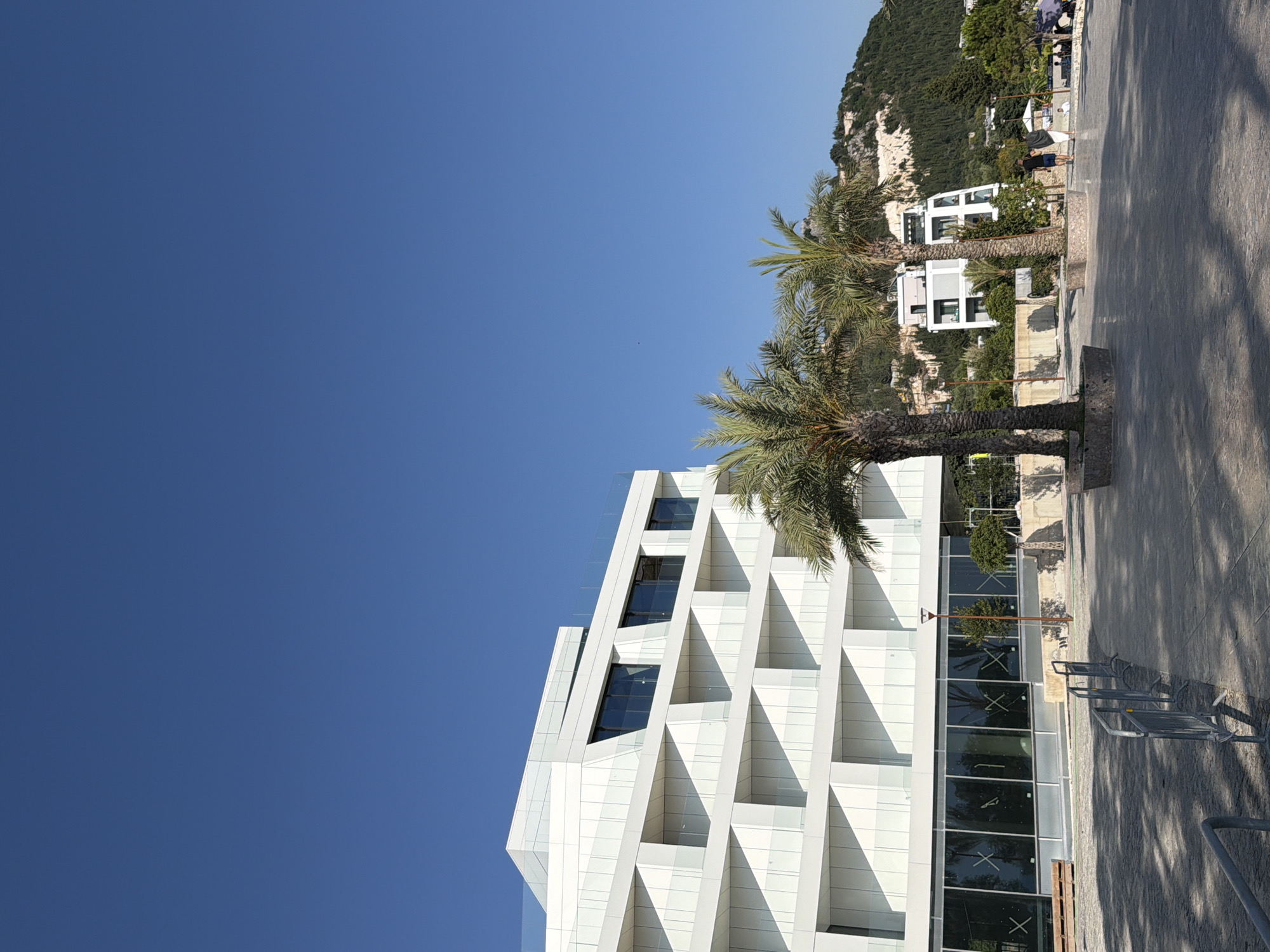 The image depicts a modern white building with geometric architectural features, situated in the city of Himarë, Albania. The building has large windows and balconies with glass railings. In front of the building, there is a paved walkway with palm trees and streetlights. In the background, a lush green hillside with scattered white structures and trees is visible under a clear blue sky. Several people are walking and standing on the walkway, and there are some temporary metal barriers in the foreground. The scene appears to be a public area, possibly a promenade or plaza, near the coast.