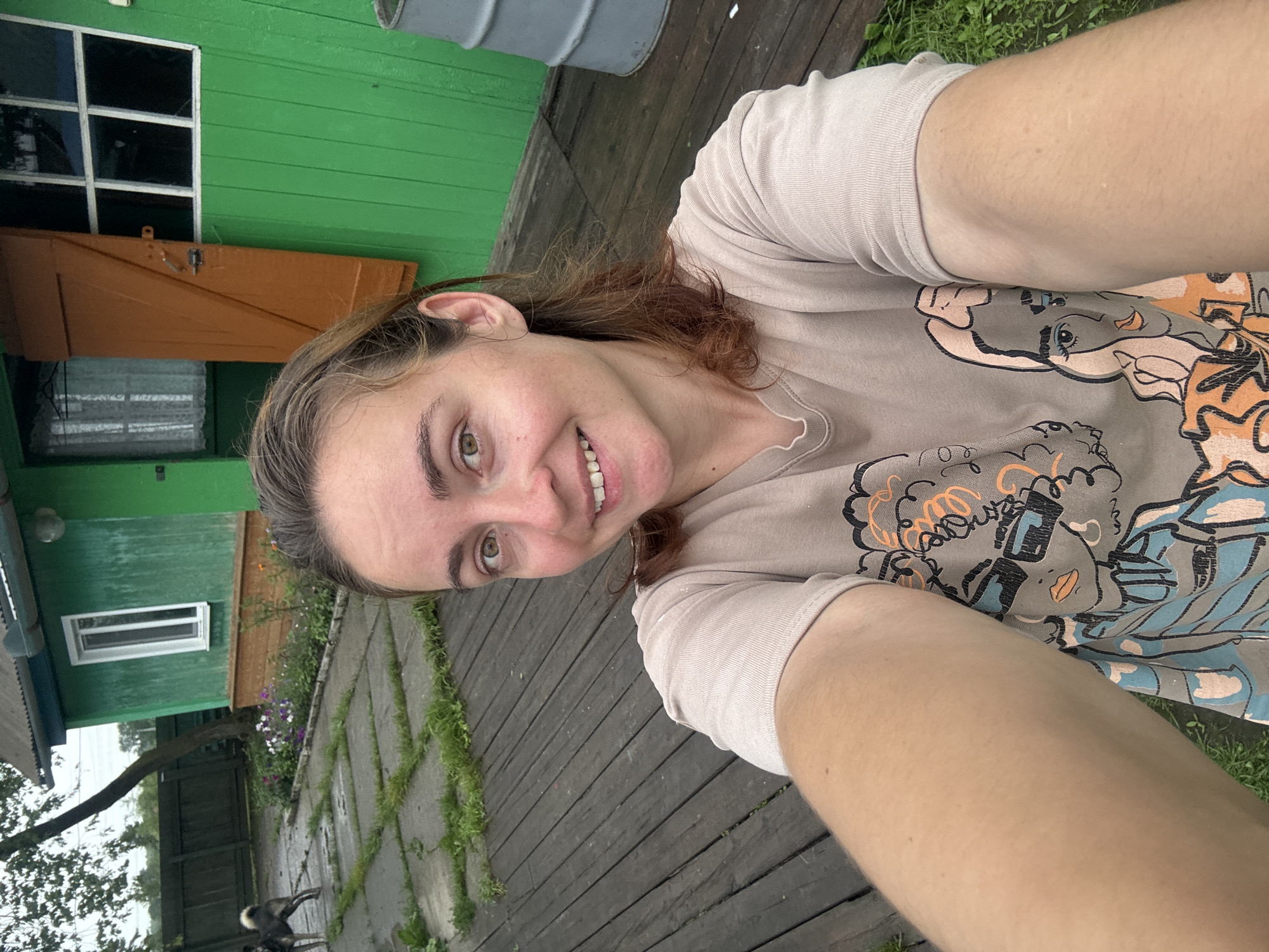 A medium-shot selfie shows a young woman with brown hair and light skin, wearing a beige t-shirt with a graphic print of two women. She is smiling at the camera. The background is a wooden deck, with a green building with orange shutters and windows behind her. There is a dog visible in the distance to the left, and a gray barrel on the right. The scene appears to be a residential yard, likely in a rural or suburban setting. The image's metadata indicates it was taken in Gonja, Russia.