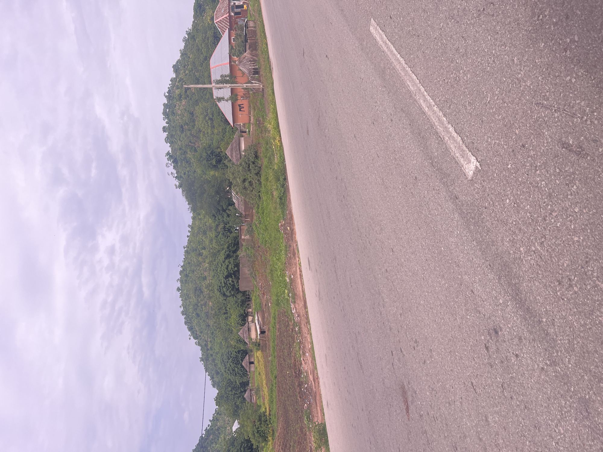 The image captures a rural landscape with a paved road in the foreground, leading into a village situated at the base of a lush, green, and slightly hilly terrain. The sky above is overcast with soft, diffused light.
In the village, several structures are visible. There are traditional huts with thatched roofs, some appearing to be made of mud. Interspersed with these are more modern-looking buildings with orange and red-tiled roofs, one of which has solar panels. A power pole stands near one of the modern buildings. The land between the road and the village appears to be cultivated with rows of young crops or cleared earth. Scattered debris can be seen along the edge of the road.
The scene suggests a serene, developing rural setting in Minda, Nigeria, where traditional and contemporary housing coexist amidst a verdant environment. There are no people or animals immediately visible in the frame.
