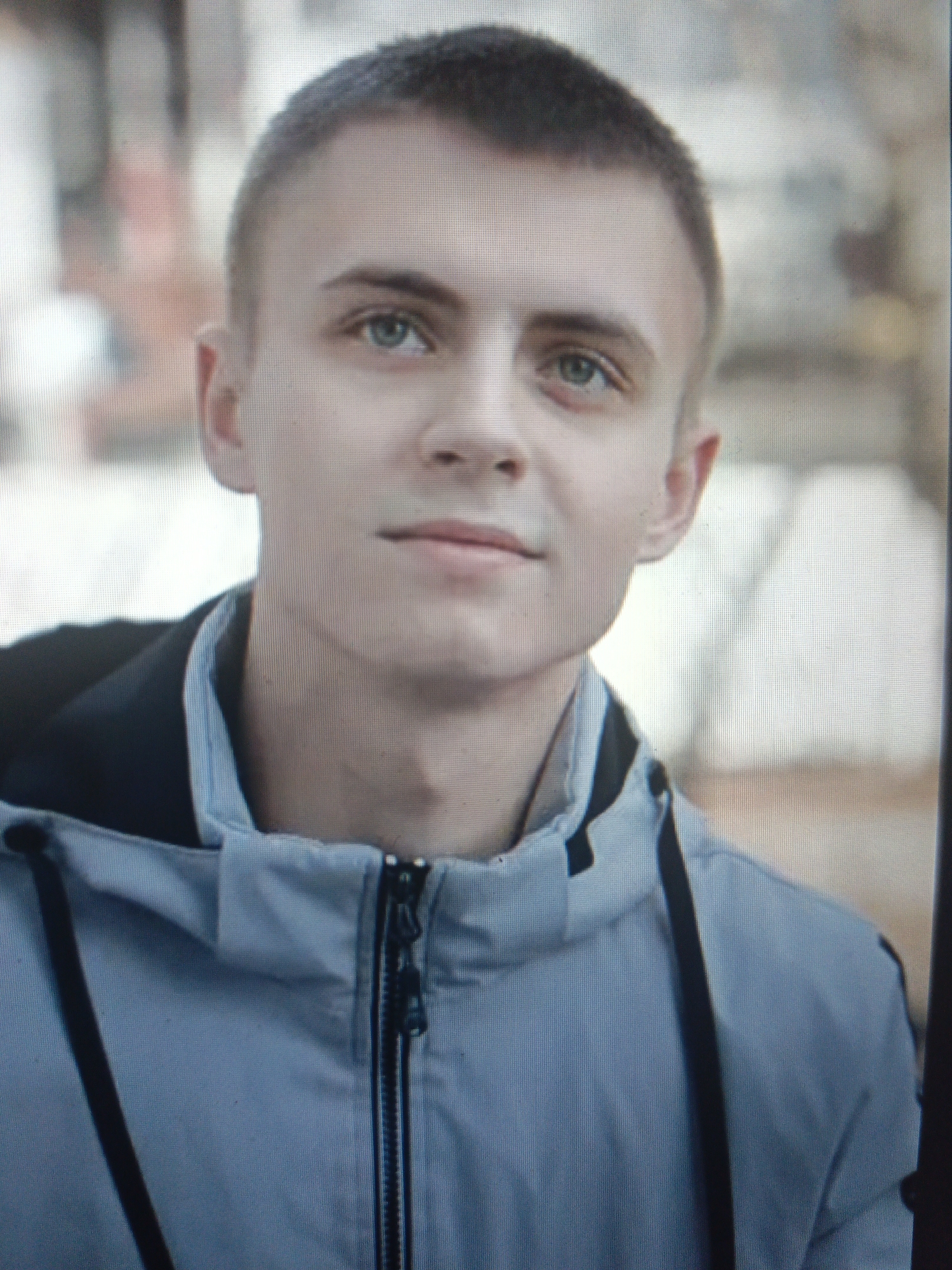 The image shows a close-up, slightly pixelated, medium shot of a young man with short, light brown hair and light blue eyes. He is wearing a light blue hooded jacket with a black zipper and black trim. The background is blurred and appears to be outdoors, with hints of foliage and buildings, suggesting an urban environment. There is no specific information about the location within the city of Uren, Russia, that can be discerned from the image.