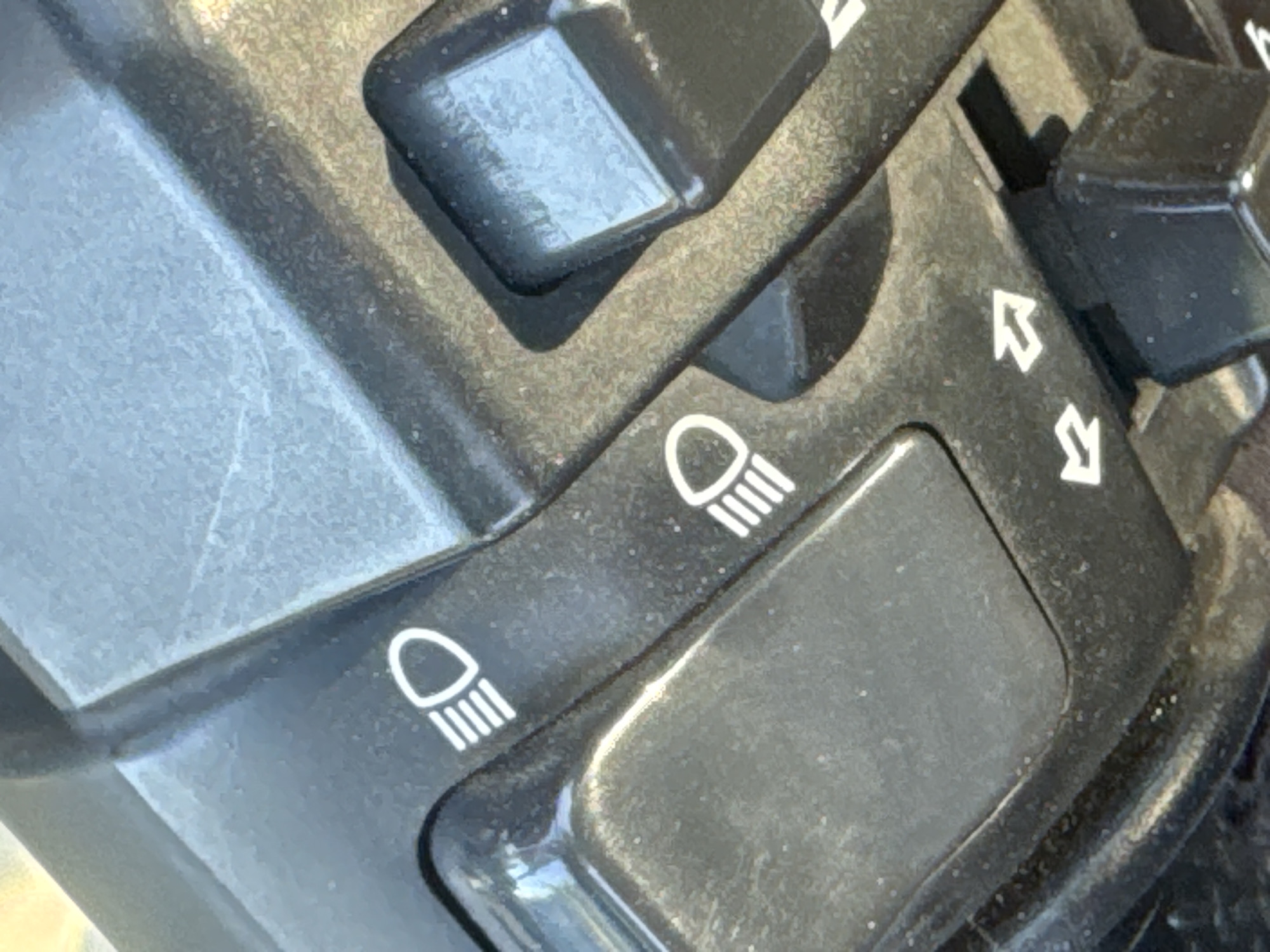 The image displays a close-up view of a black control panel, likely from a vehicle. Two icons of a headlight are visible, one above the other and slightly to the left. These icons are white and consist of a semicircular shape with three vertical lines emanating from the bottom. Between and slightly above the headlight icons, there are two white arrow symbols pointing upwards. The panel itself appears to be made of plastic with some textured areas and a smooth, glossy section. The lighting suggests it is daytime, but there are no clear indications of weather or a specific outdoor location beyond the general context of a vehicle. No people are visible in the image. The overall scene is static, focusing on the detailed textures and symbols of the control panel. The setting appears to be outdoors given the lighting and the close-up focus on what seems to be part of a motorcycle or scooter.