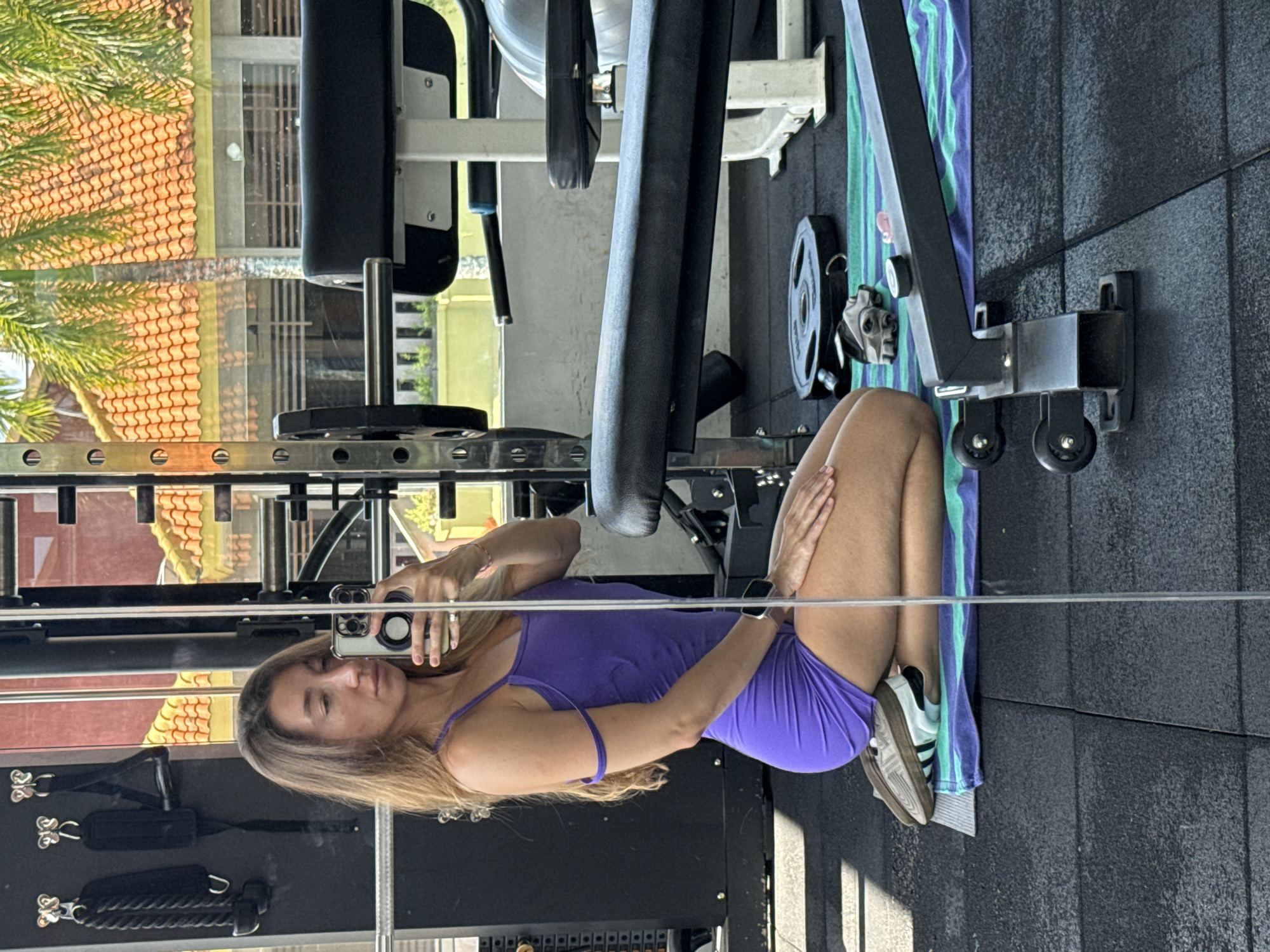 A woman in a purple dress and sneakers is kneeling on a mat in a gym. She is taking a selfie in a mirror. The gym appears to be located in Kuta Selatan, Indonesia, as evidenced by the distinct architecture and vegetation visible through the gym's windows. The background features palm trees, terracotta-tiled roofs, and a building with a yellow facade, characteristic of the region. Gym equipment, including weight plates, gloves, and a weight bench, are visible in the foreground and background. The flooring is a black rubber gym mat.