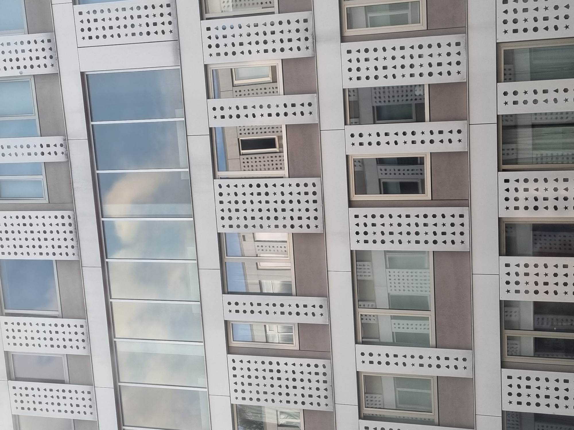 The image shows a close-up of a modern building facade. The building has a repetitive pattern of rectangular windows alternating with gray panels that feature a regular pattern of cutout shapes: stars, circles, hexagons, and triangles. The windows on the left reflect the sky and clouds. The building appears to be located in a city, but the specific location is not evident.